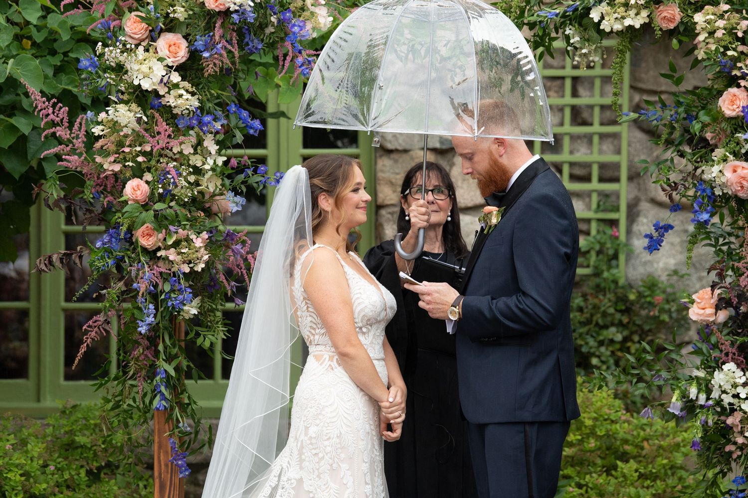 Rainy wedding ceremony under clear umbrellas with floral arch and officiant.