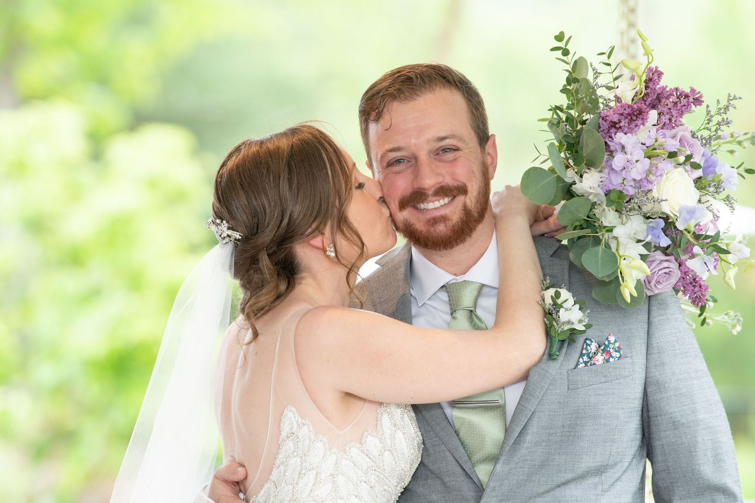 A joyful wedding couple shares an intimate moment outdoors with purple floral decorations in the background.