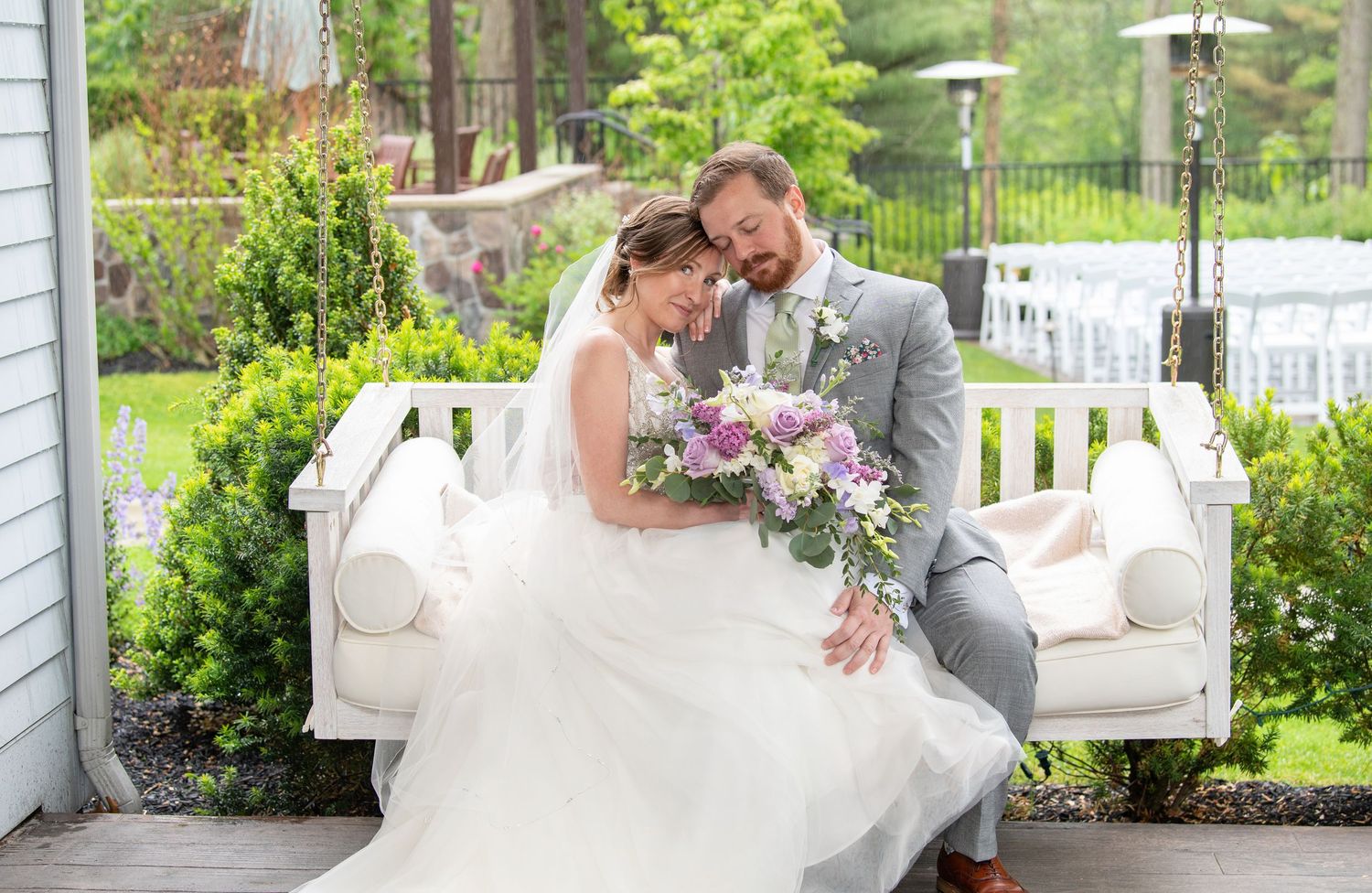 A couple in wedding attire sits together on a white porch swing surrounded by lush greenery.