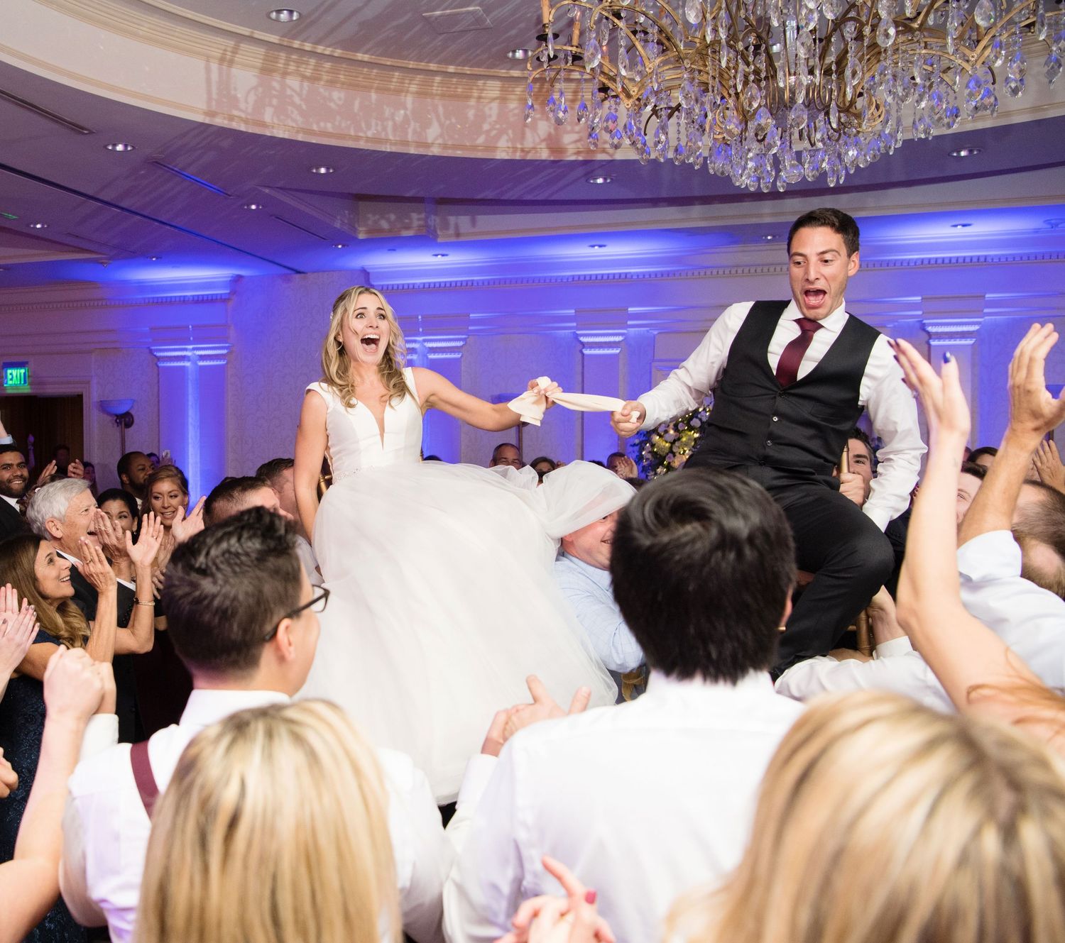 Wedding guests on a dance floor celebrating with hands raised beneath elegant chandeliers and blue lighting.