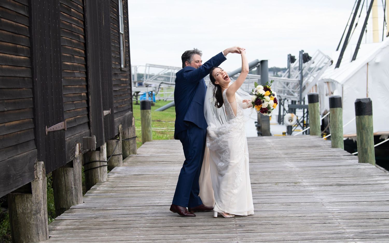 A couple in formal attire stands on a wooden boardwalk embracing by the waterfront as their photos are being taken.