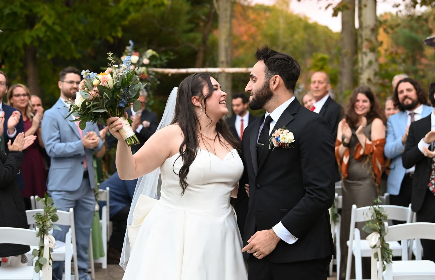 A joyful outdoor wedding ceremony with guests looking on as a couple celebrates at the altar with white chairs and greenery.