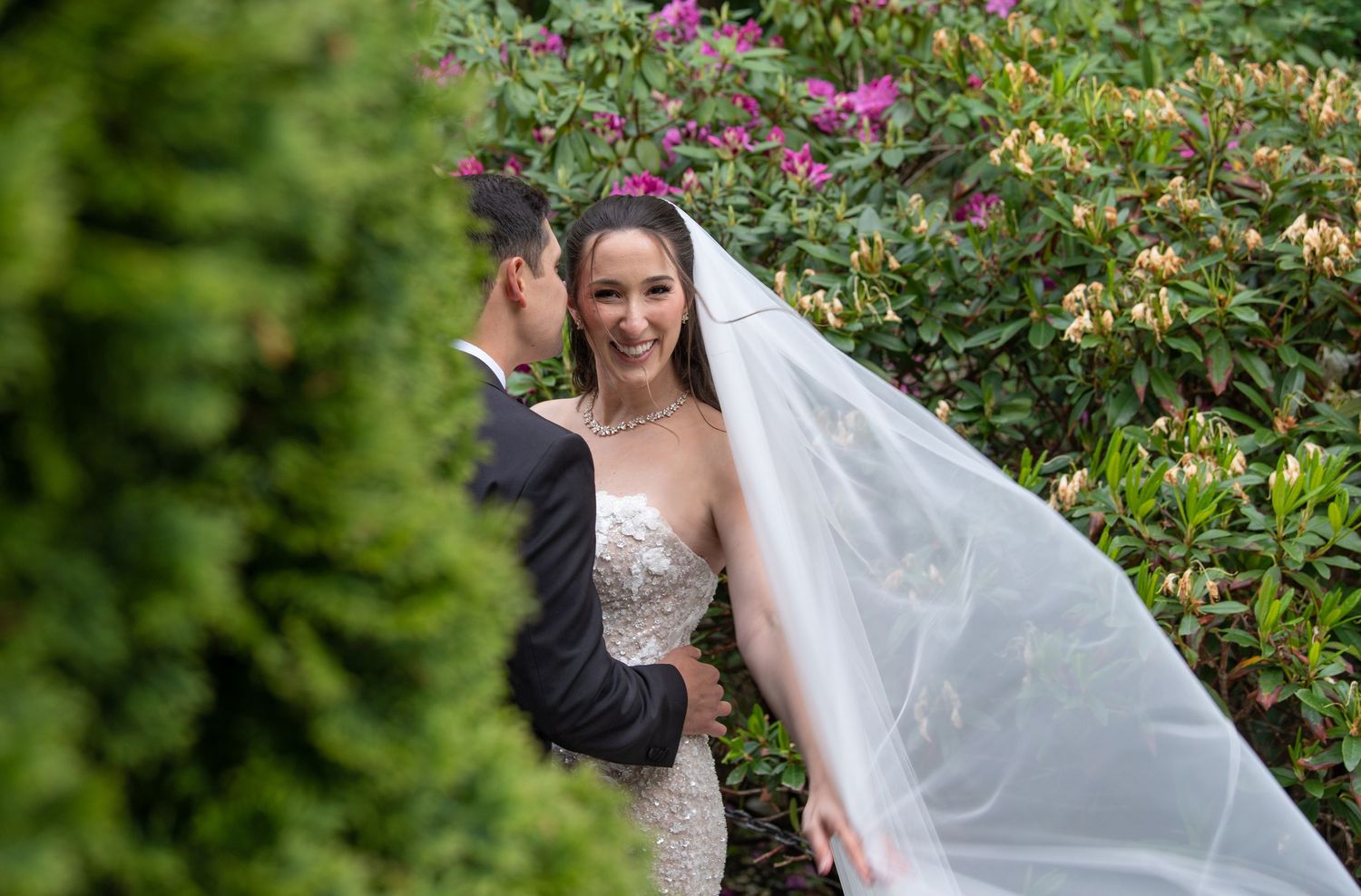 Bride in strapless gown with flowing veil stands beside groom in tuxedo among lush green hedges and pink blooms.