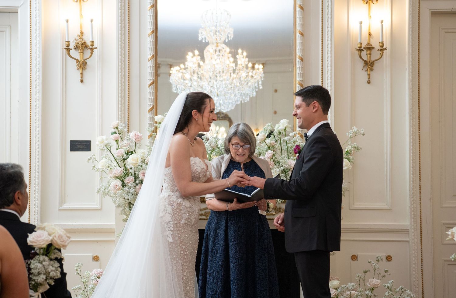 Bride and groom exchanging vows at elegant indoor wedding ceremony with chandelier and floral arrangements.