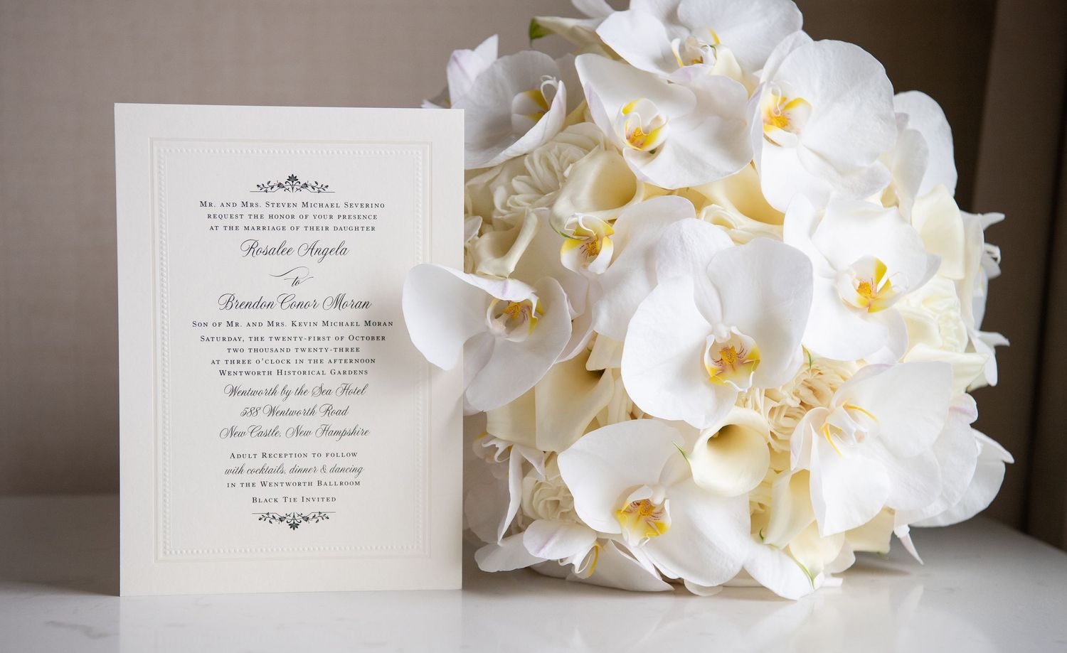 An elegant white wedding invitation sits beside a luxurious bouquet of white phalaenopsis orchids.