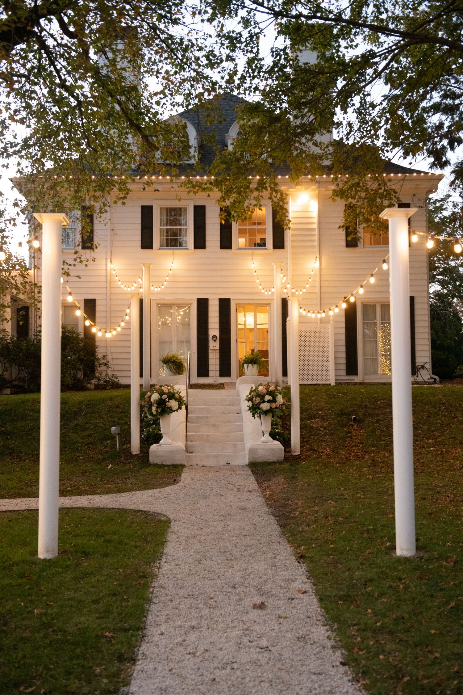 White pillared mansion with string lights illuminating a curved gravel pathway at dusk.