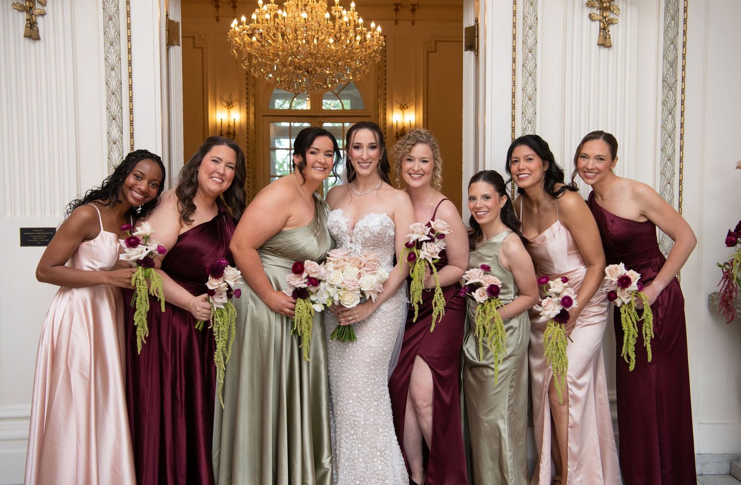 Bride in white gown poses with seven bridesmaids in burgundy and blush dresses holding floral bouquets in elegant ballroom.