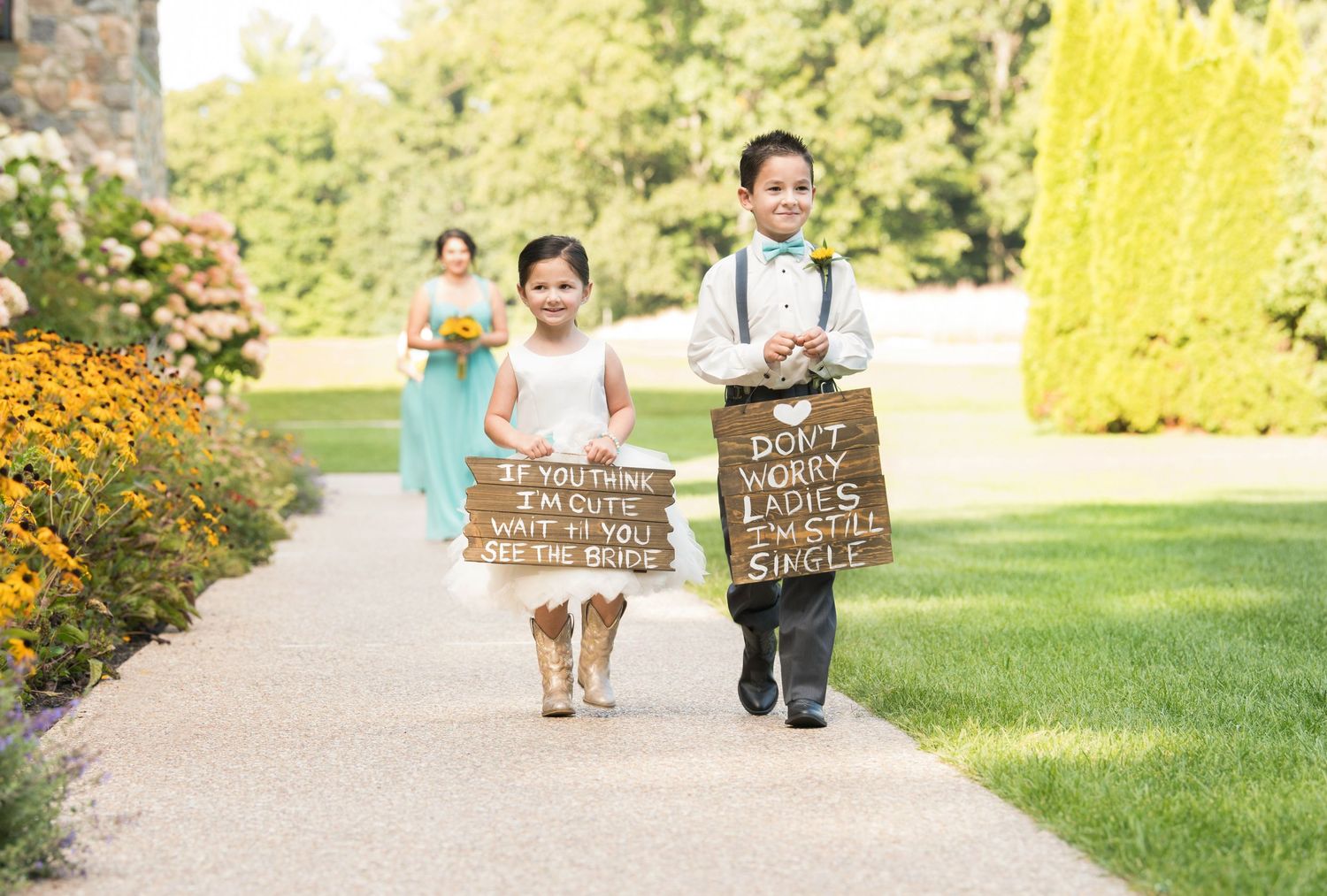 Children walking down garden path carrying rustic wooden signs at outdoor wedding ceremony.