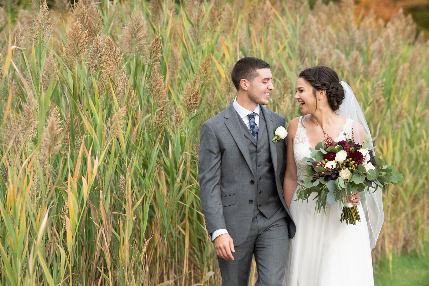 A couple in wedding attire shares a joyful moment together in a field of tall grass during a beautiful autumn day.