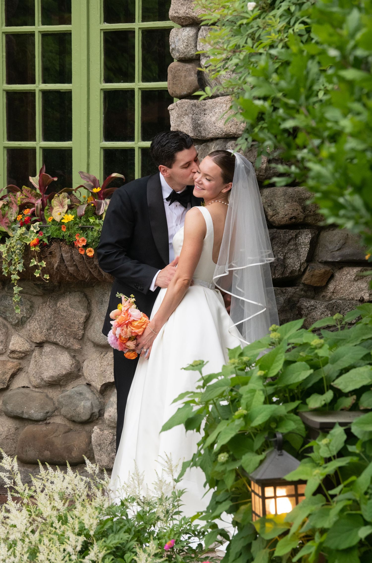 Bride and groom share a romantic kiss in a lush garden, bride holding a vibrant orange bouquet in a white gown.