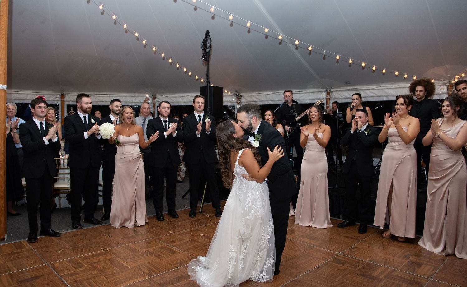 Wedding guests dance together in a reception hall decorated with string lights and neutral decor.