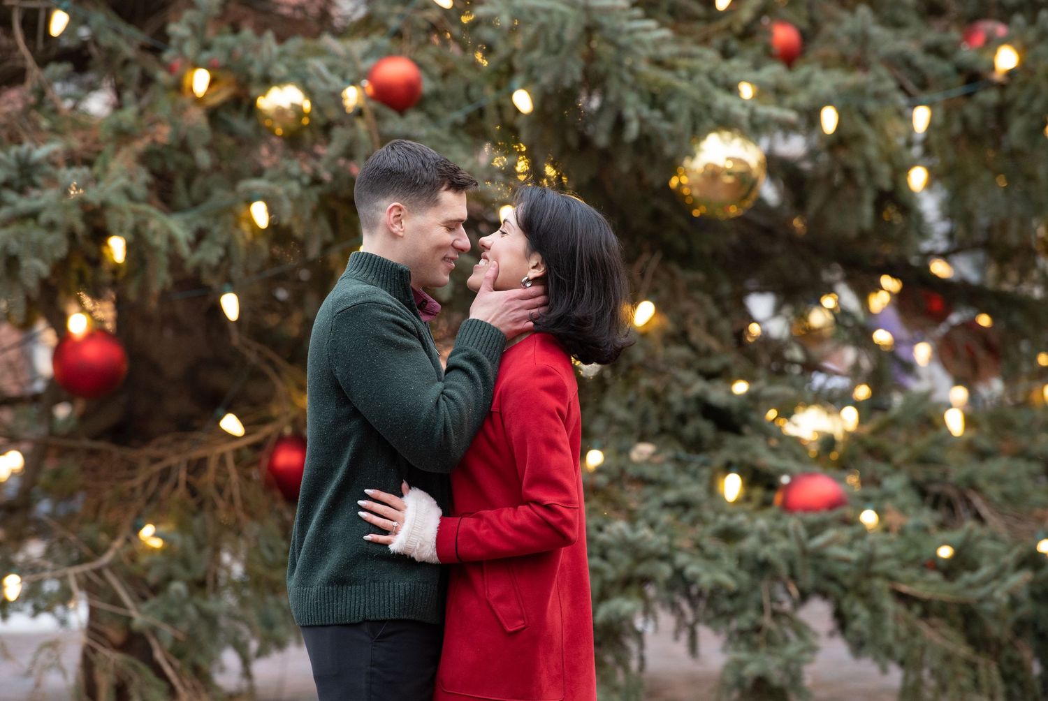 A couple in winter attire embraces beneath twinkling holiday lights and decorated evergreen trees.