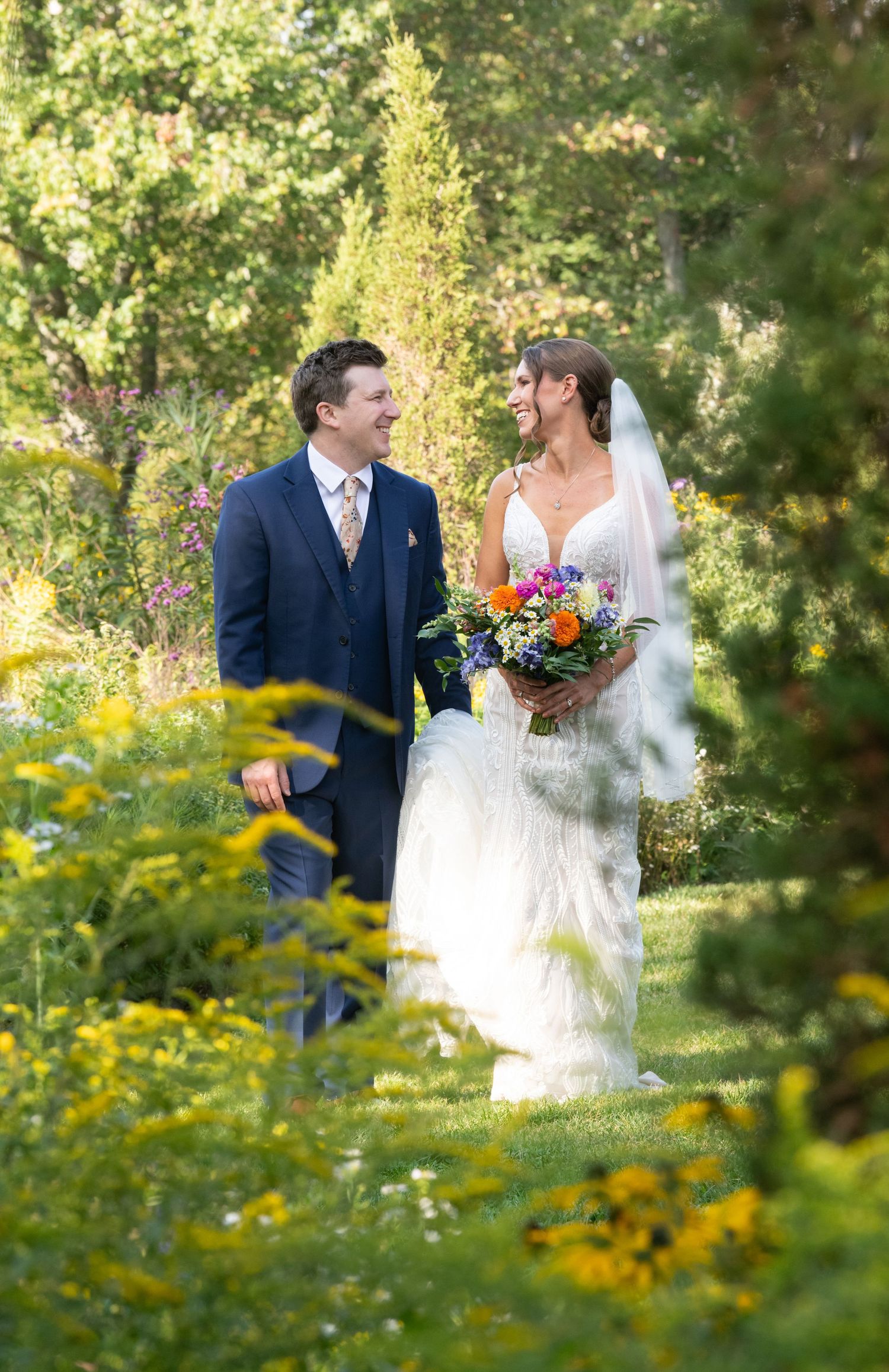 A smiling bride and groom walk through a lush garden, she holds a colorful bouquet and wears a white gown with veil.