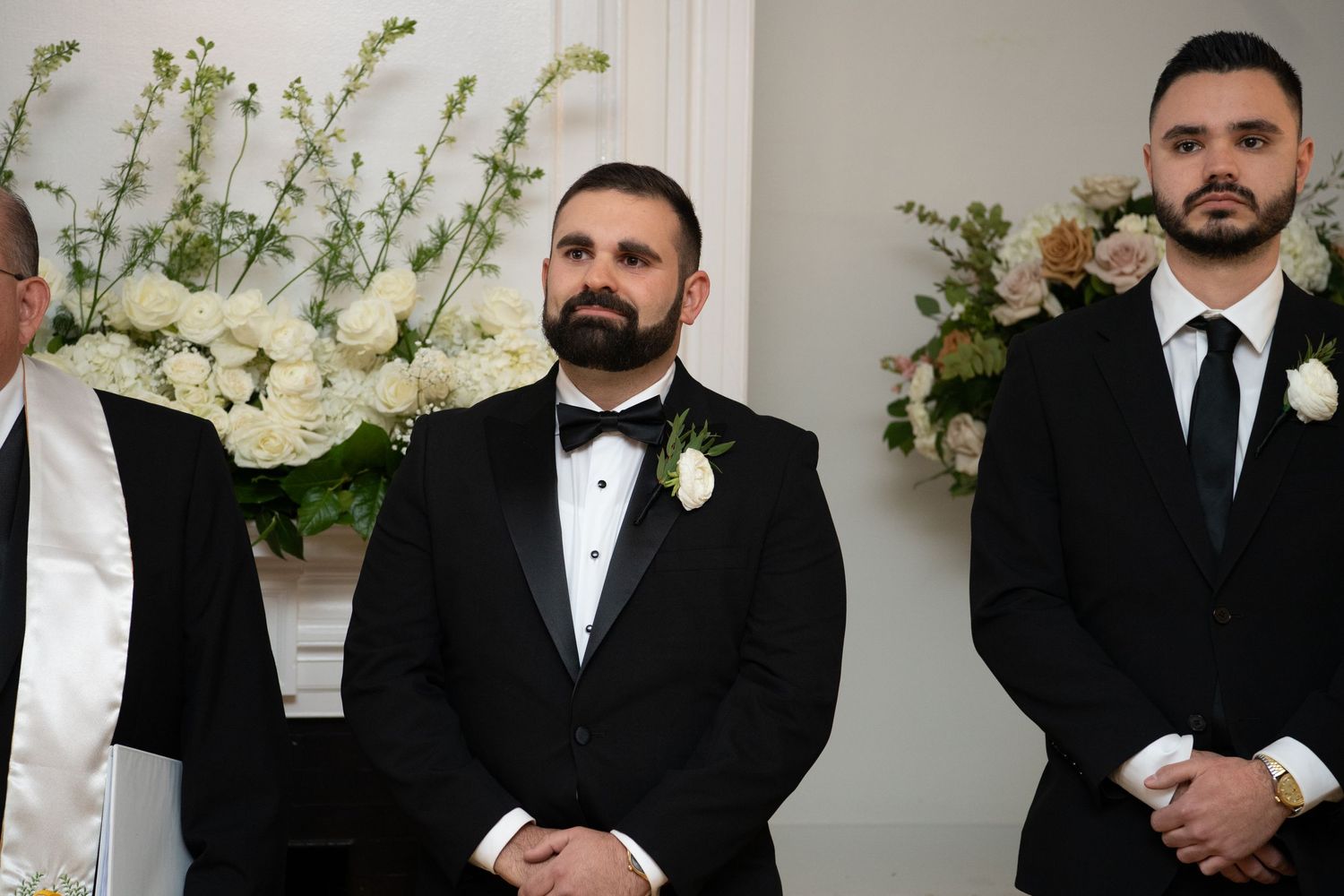 Groomsmen in black tuxedos and white shirts stand in a line against a white wall decorated with floral arrangements.