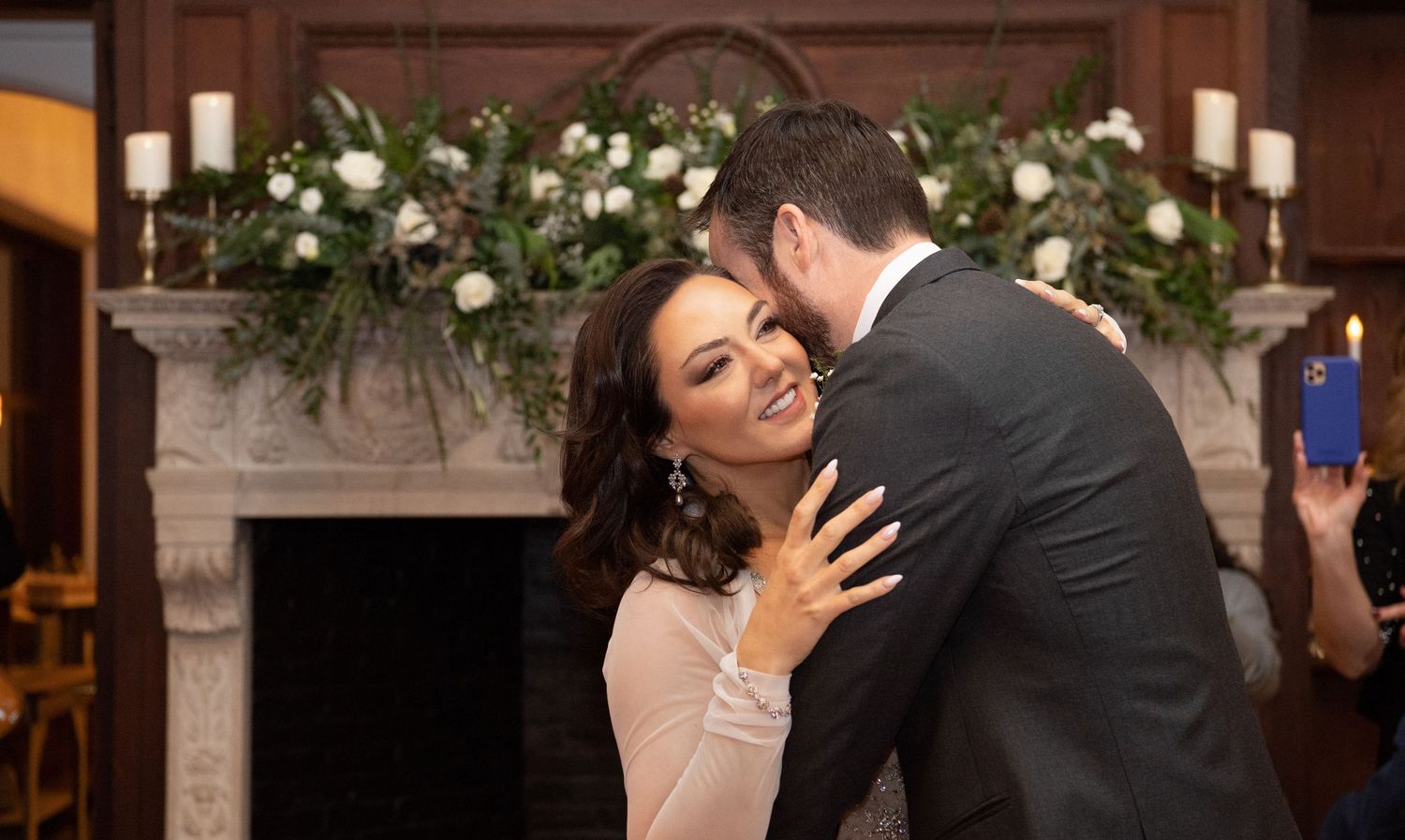 A romantic moment captured during a wedding celebration in front of a fireplace decorated with white floral garlands.