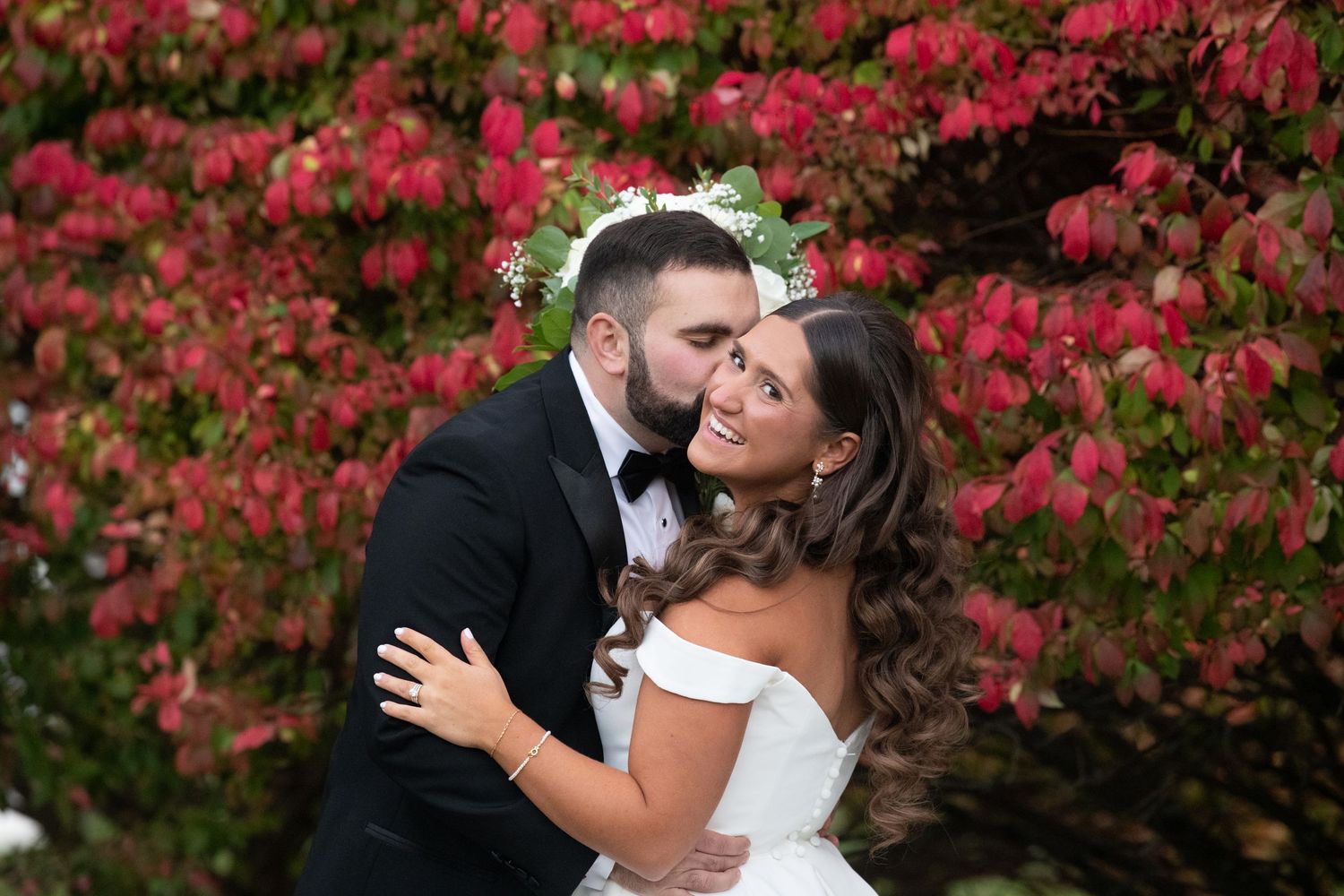A romantic wedding couple embraces in front of vibrant red flowering bushes during their outdoor photo session.