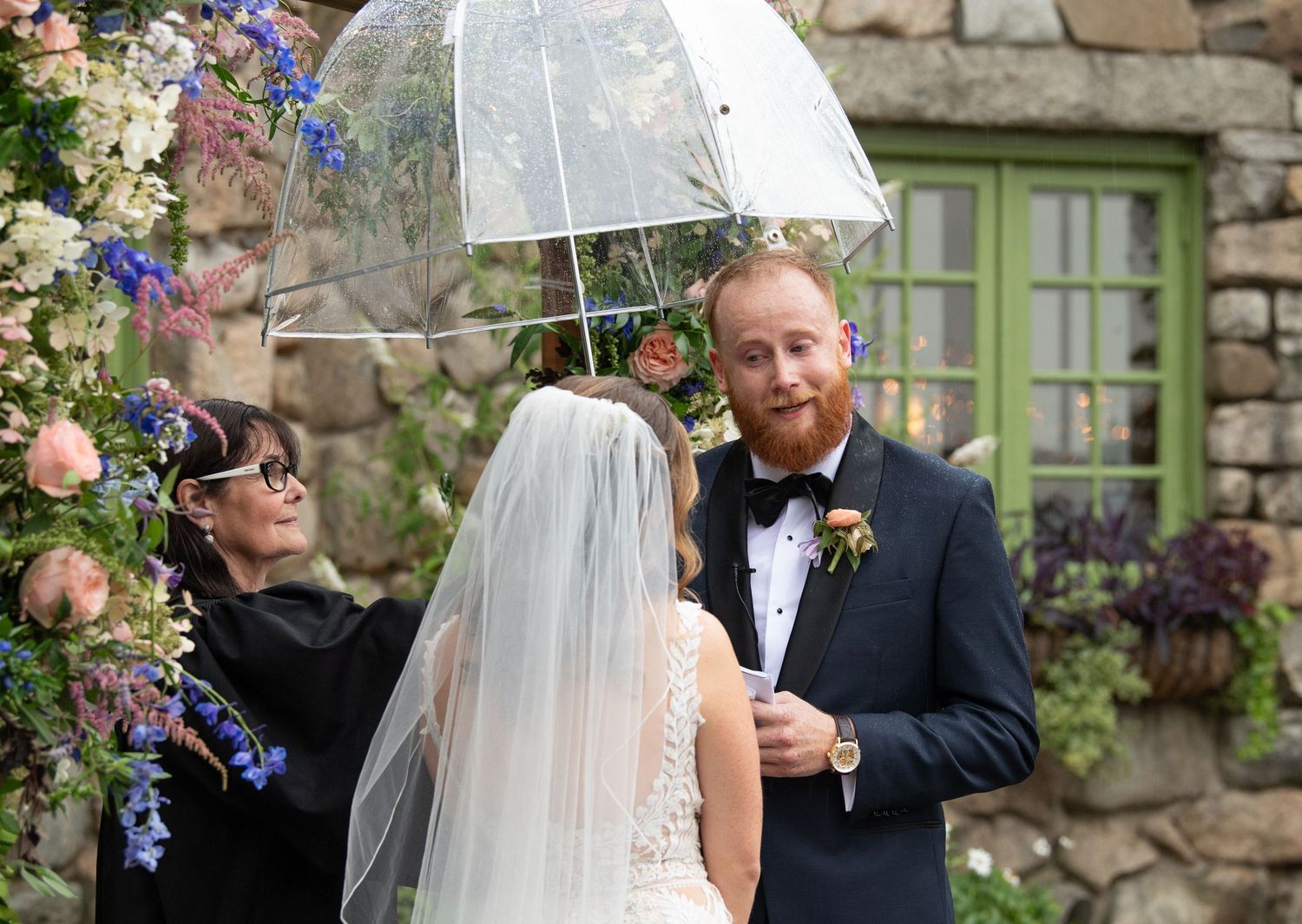 A wedding ceremony takes place under clear umbrellas with colorful floral arrangements against stone architecture.