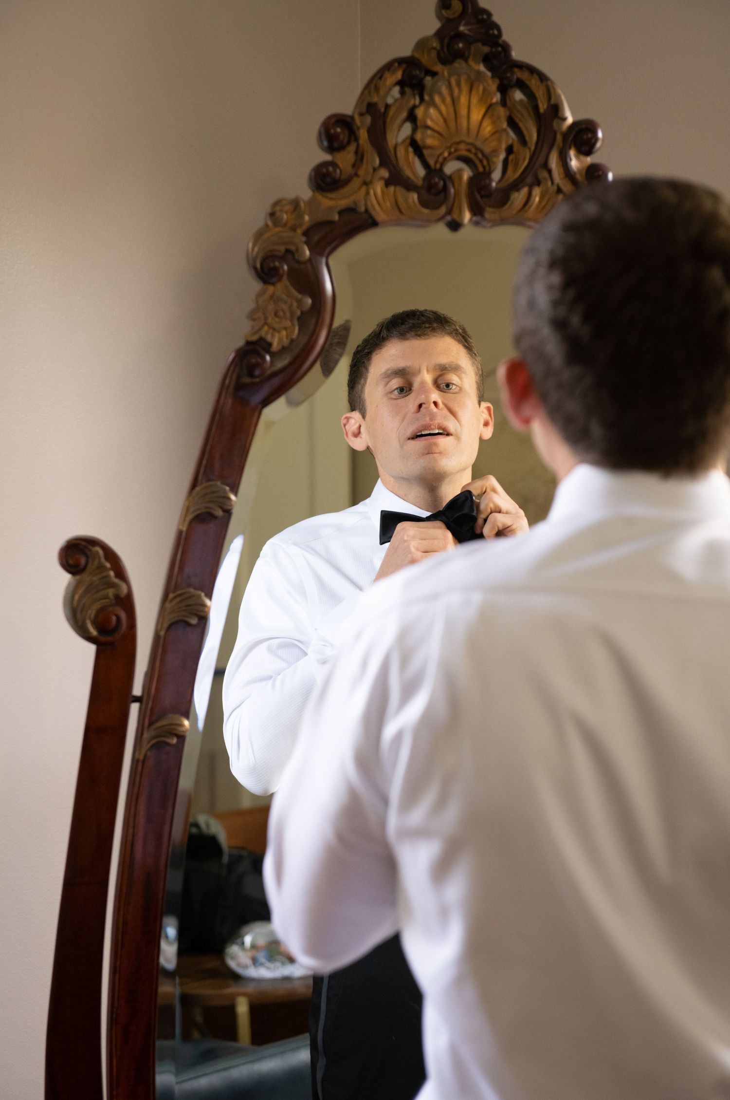 Someone in formal attire fixes their tie while getting ready in front of a decorative wall mirror.