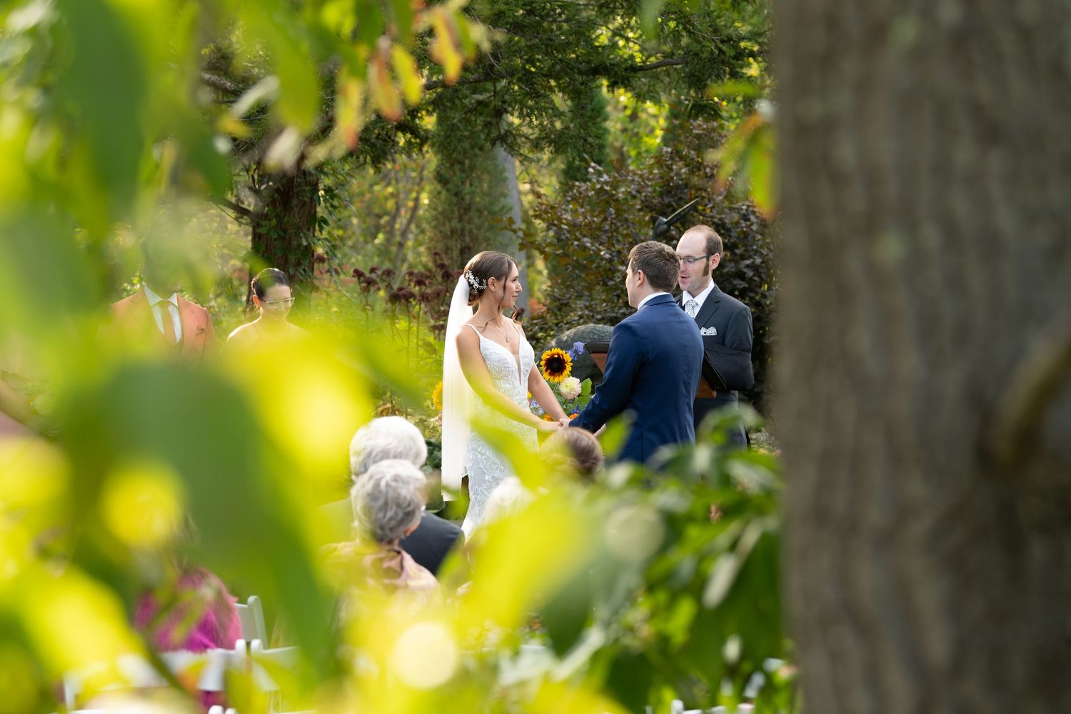 Wedding guests mingle outdoors under trees with soft natural lighting creating a romantic garden atmosphere.
