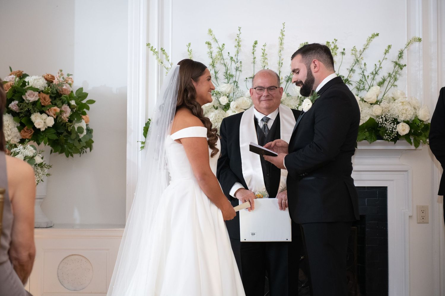 Wedding ceremony in progress in an elegant white room with decorative floral arrangements.