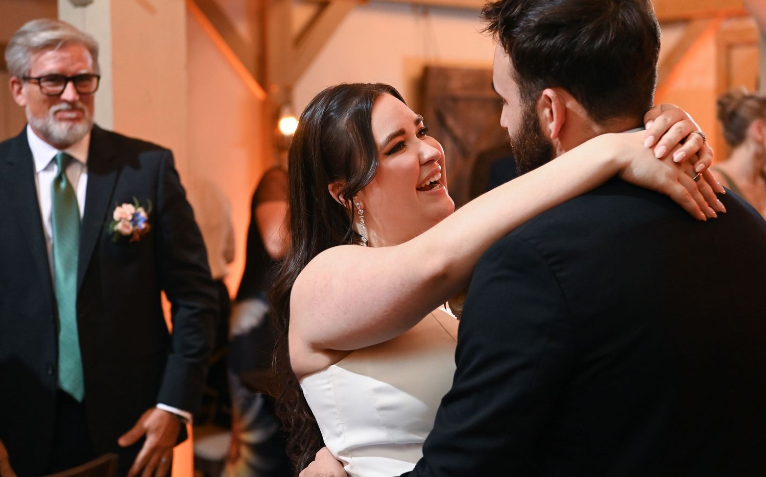 A joyful couple embraces on their wedding dance floor while an older guest in a green tie watches in the background.