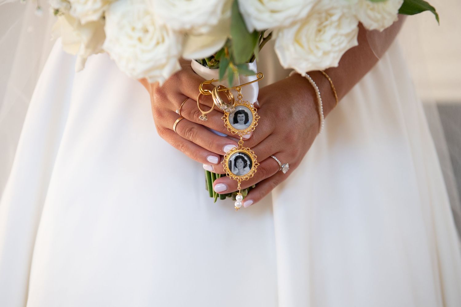 Hands adorned with gold rings hold a white rose bouquet against a flowing white wedding dress.