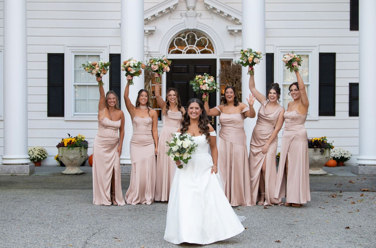 A bridal party poses outside a white colonial-style building wearing blush pink bridesmaid dresses and a white wedding gown.