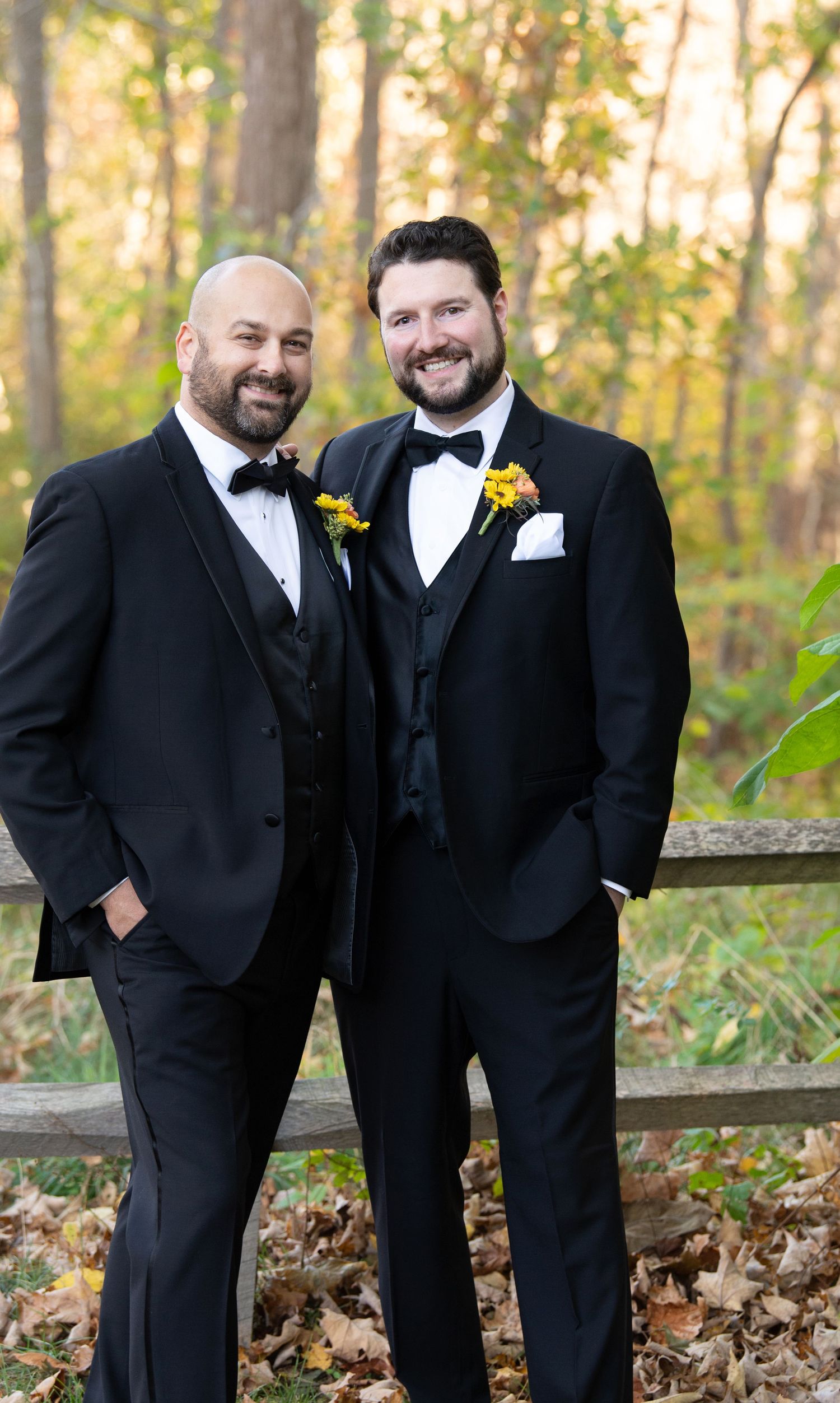 Groomsmen in black tuxedos and bowties pose together on a wooden fence during autumn with fallen leaves.