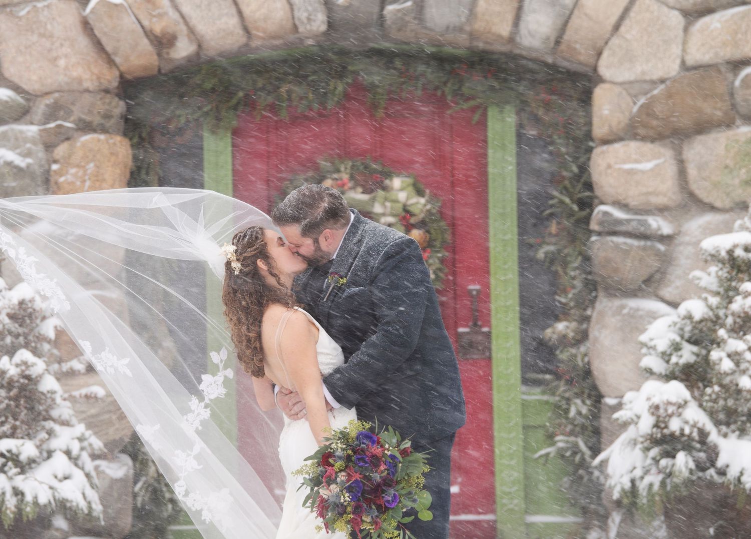 Winter wedding portrait with couple kissing in front of red door with snow falling. Willowdale Estate wedding