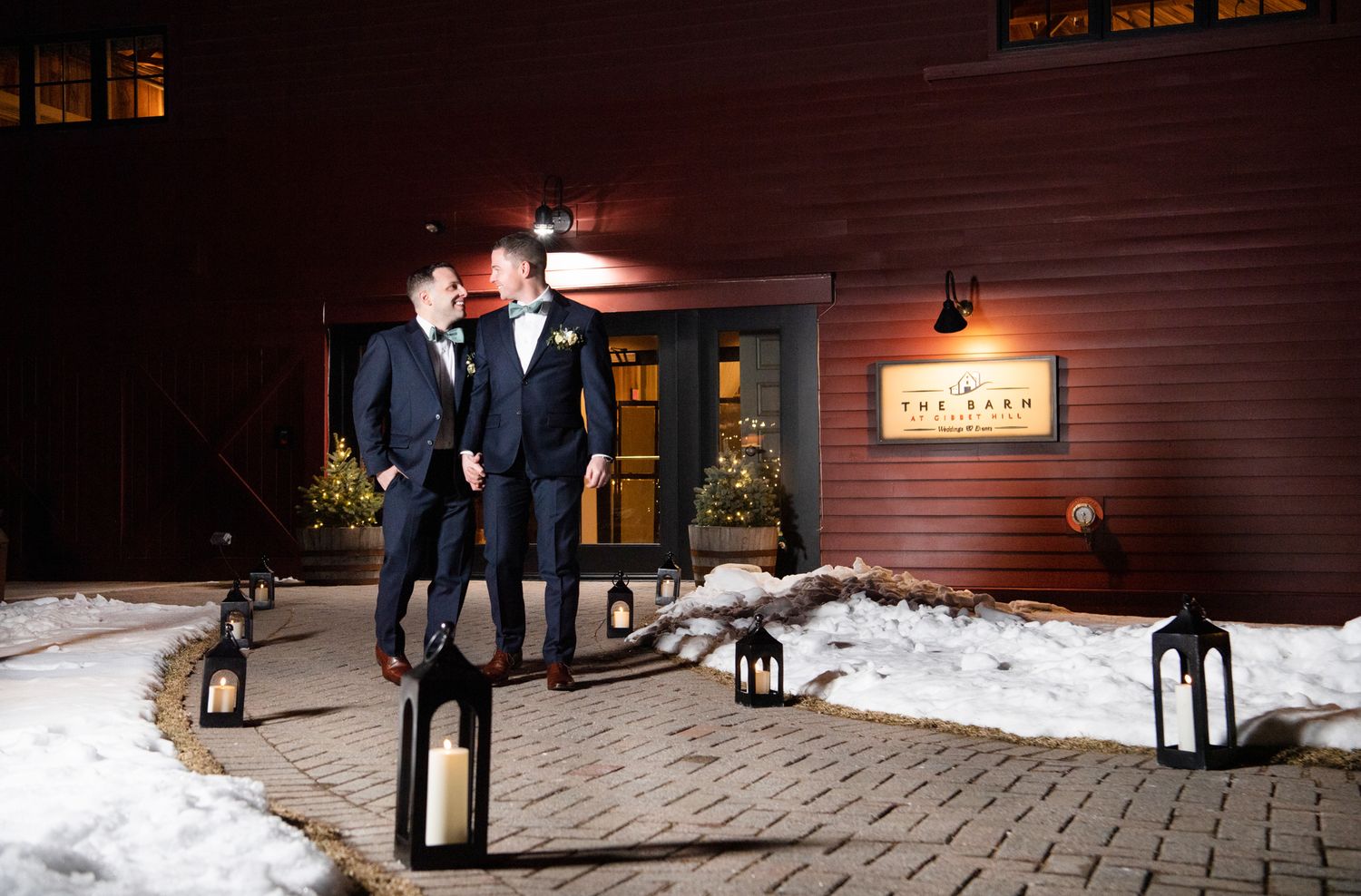Black lanterns illuminate a snow-covered pathway outside a rustic wedding venue at night.