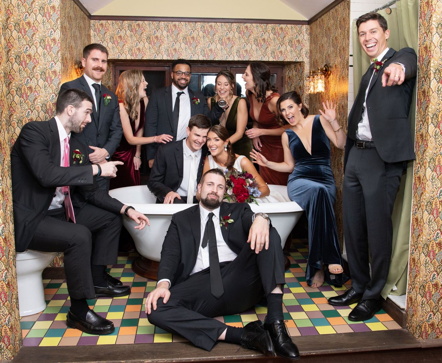 Wedding party members in formal attire pose playfully on a patterned floor in an elegant room with vintage wallpaper.