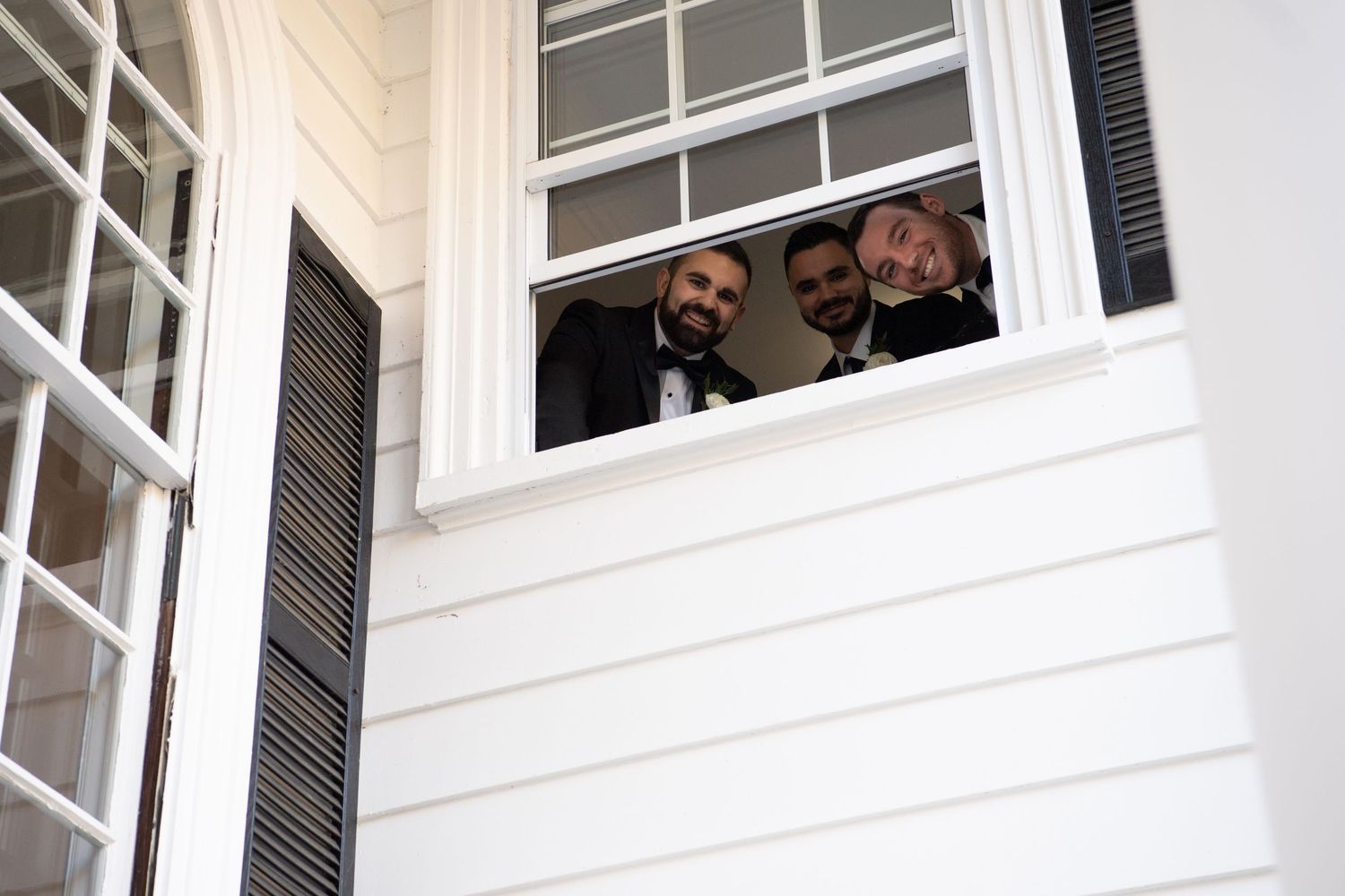 People smile and wave from windows of a white building with traditional shutters and architectural details.