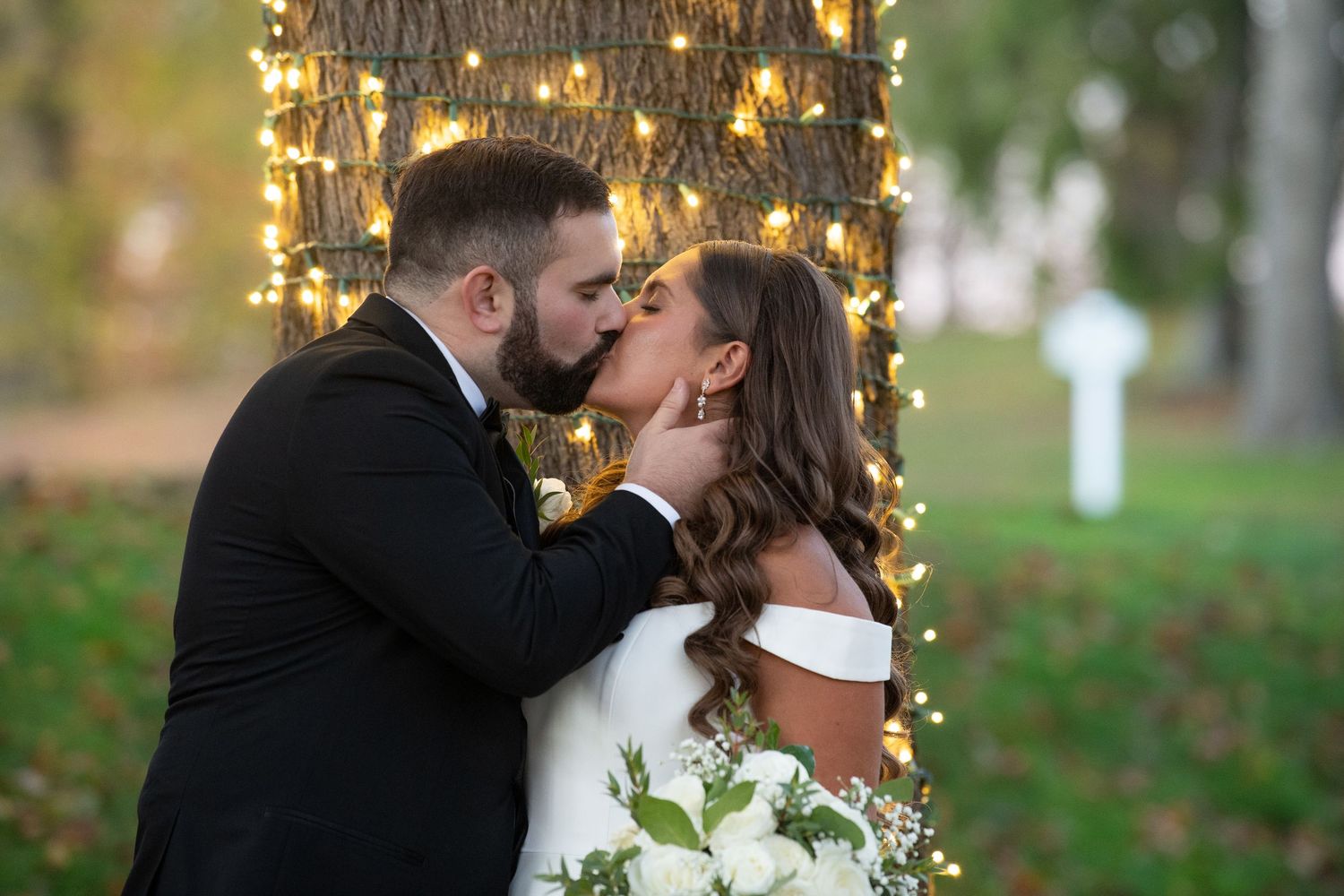 A romantic wedding couple shares a kiss outdoors with twinkling string lights and autumn leaves in the background.
