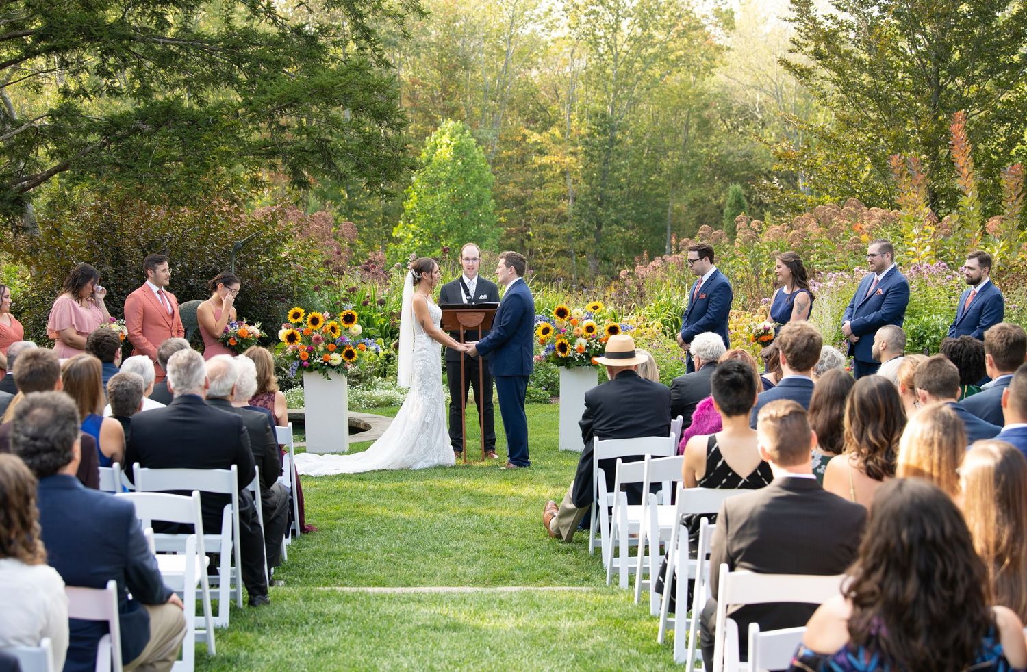 Outdoor wedding ceremony with couple at sunflower-adorned altar, guests seated on white chairs, surrounded by lush green gardens.