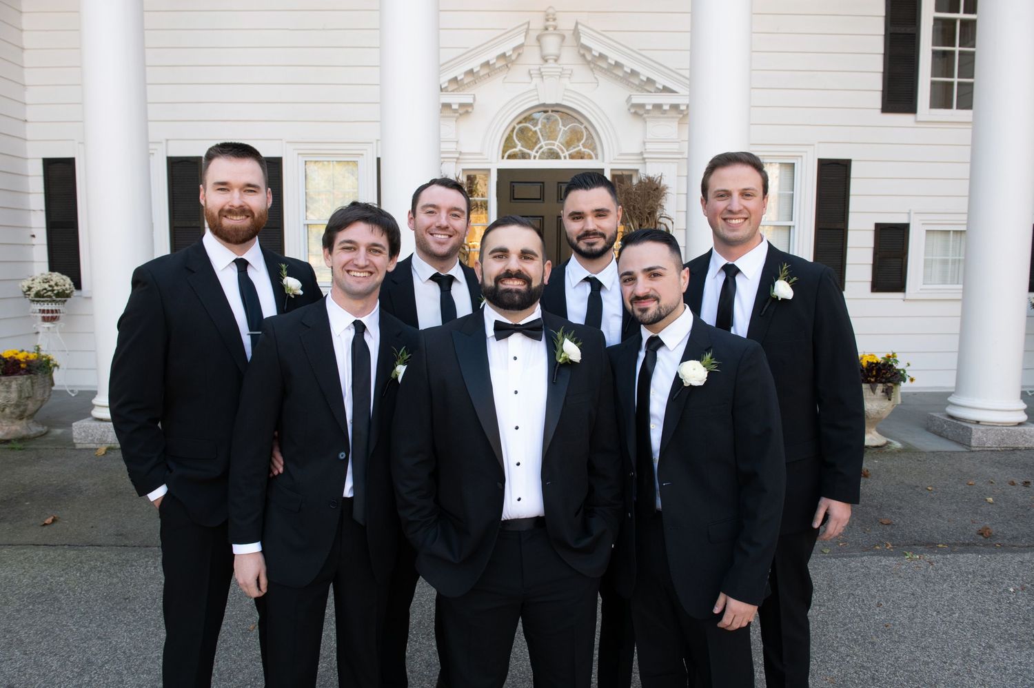 A group of formally dressed men in black tuxedos and bow ties stand together smiling outside a white building.