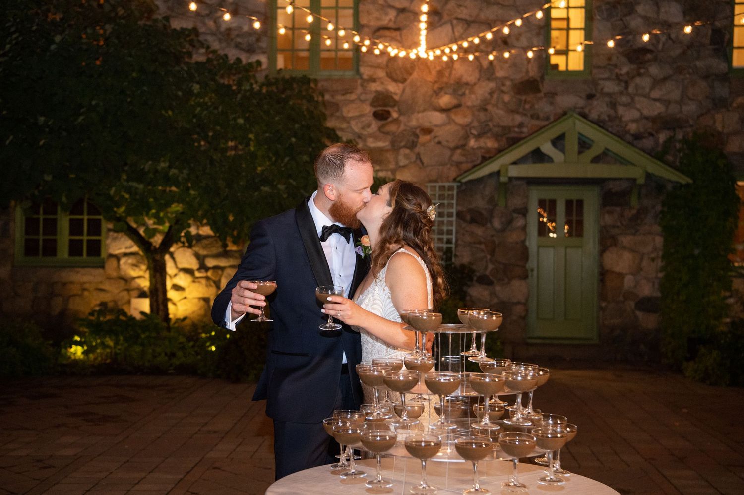 Evening wedding reception with string lights illuminating stone wall backdrop.