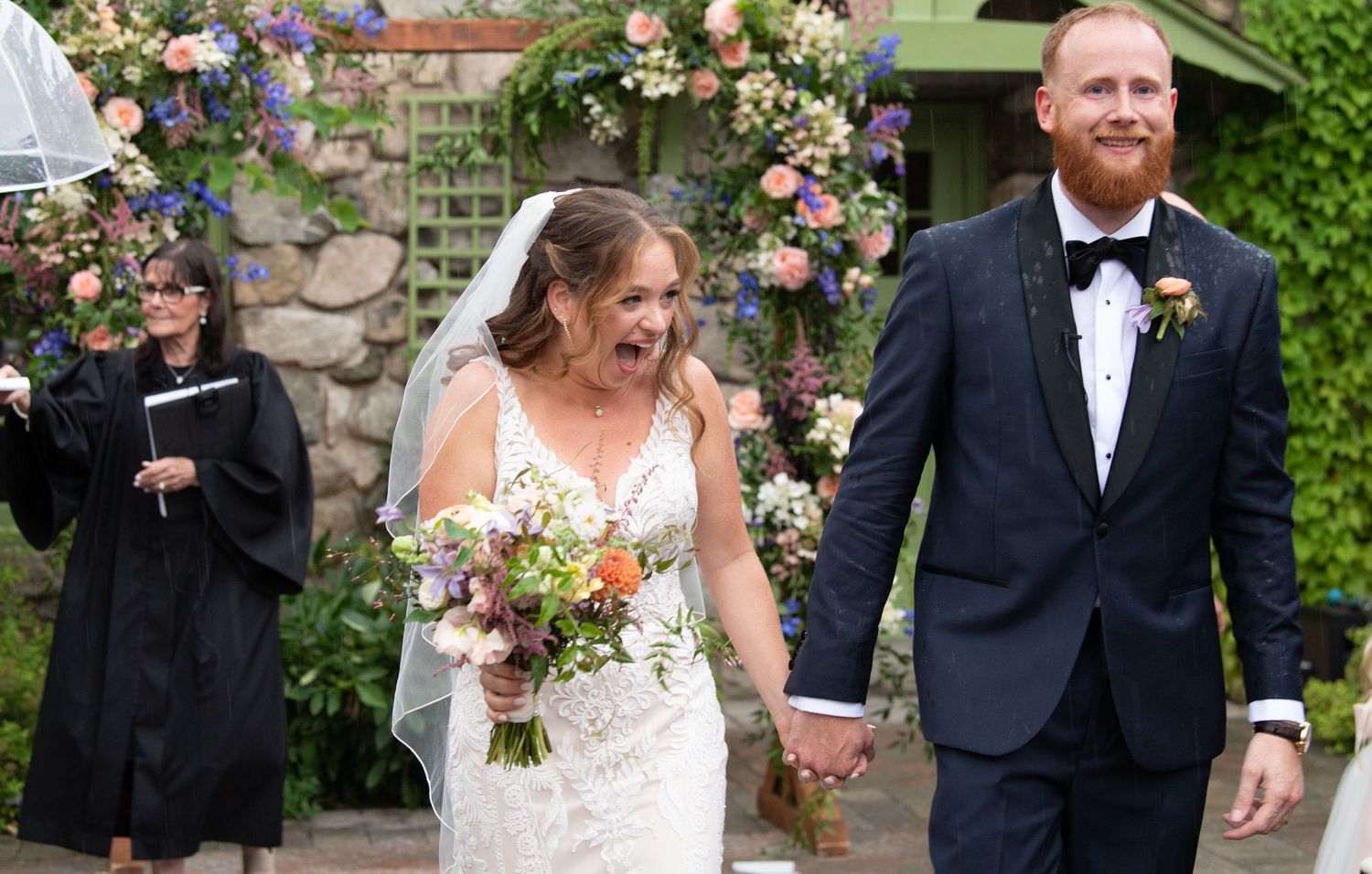 Wedding couple walks under floral arch decorations during outdoor garden ceremony.