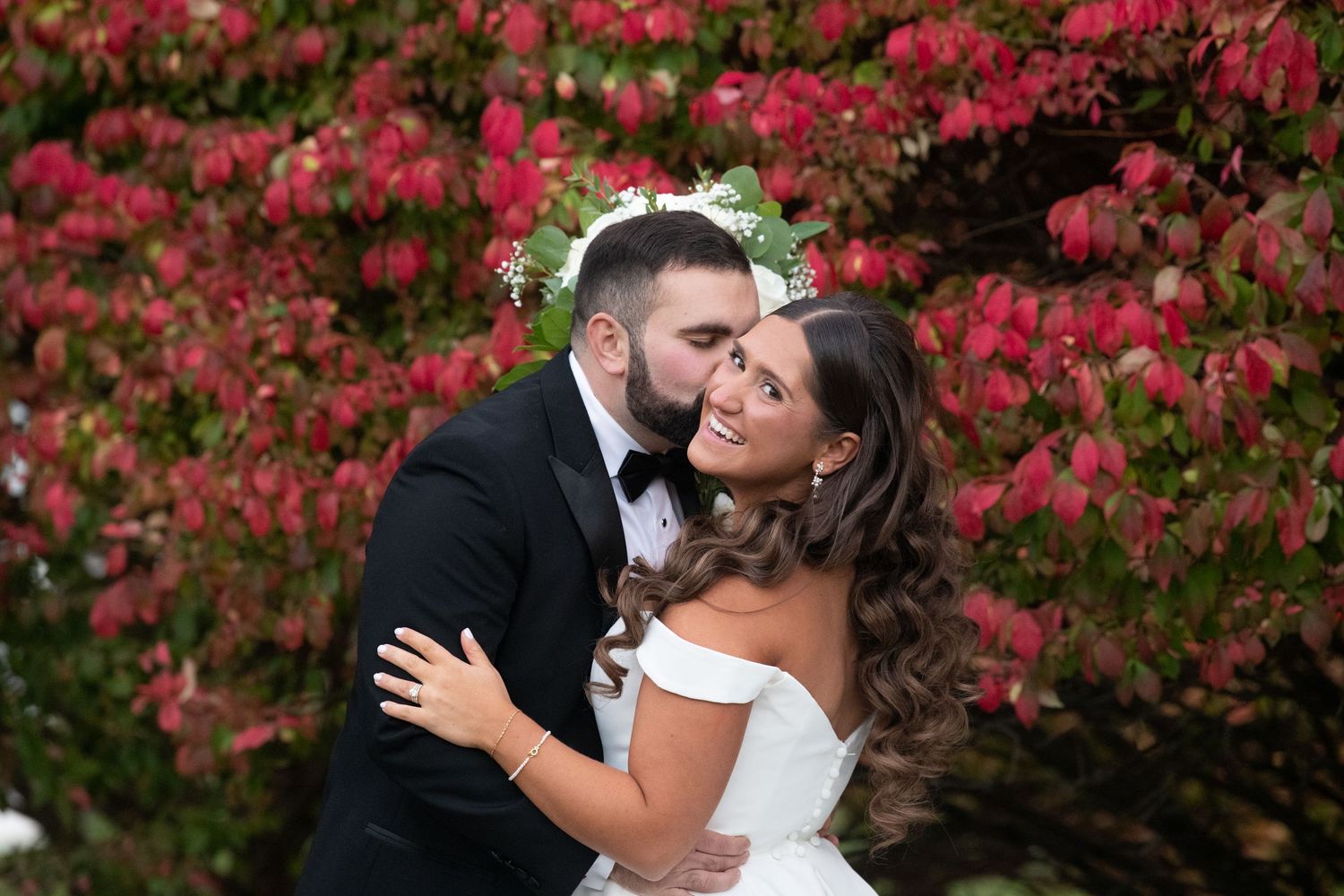 A romantic wedding couple embraces in front of vibrant red autumn foliage during their outdoor photo session.