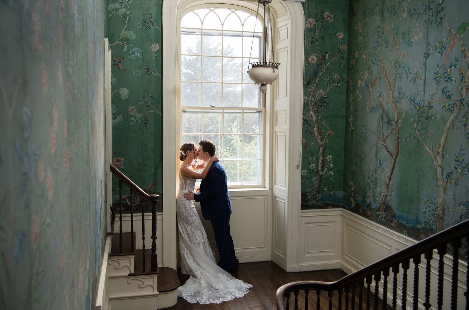 A couple in wedding attire stands in an elegant room with distressed turquoise walls and ornate windows.