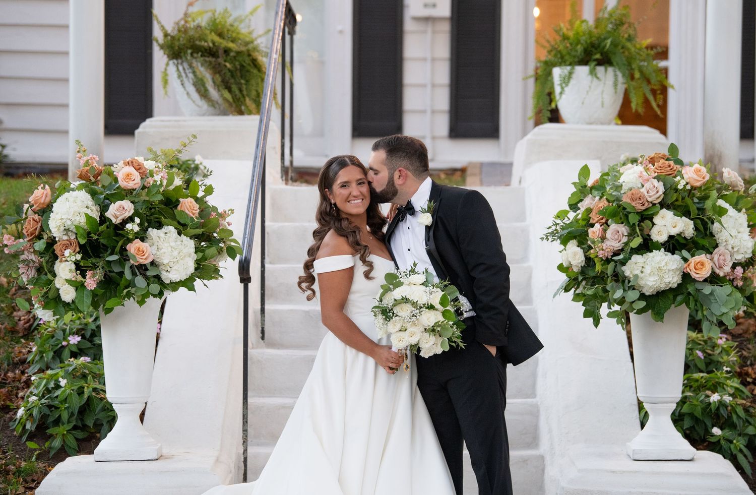 Elegant wedding couple poses together in front of a white building with beautiful floral arrangements.