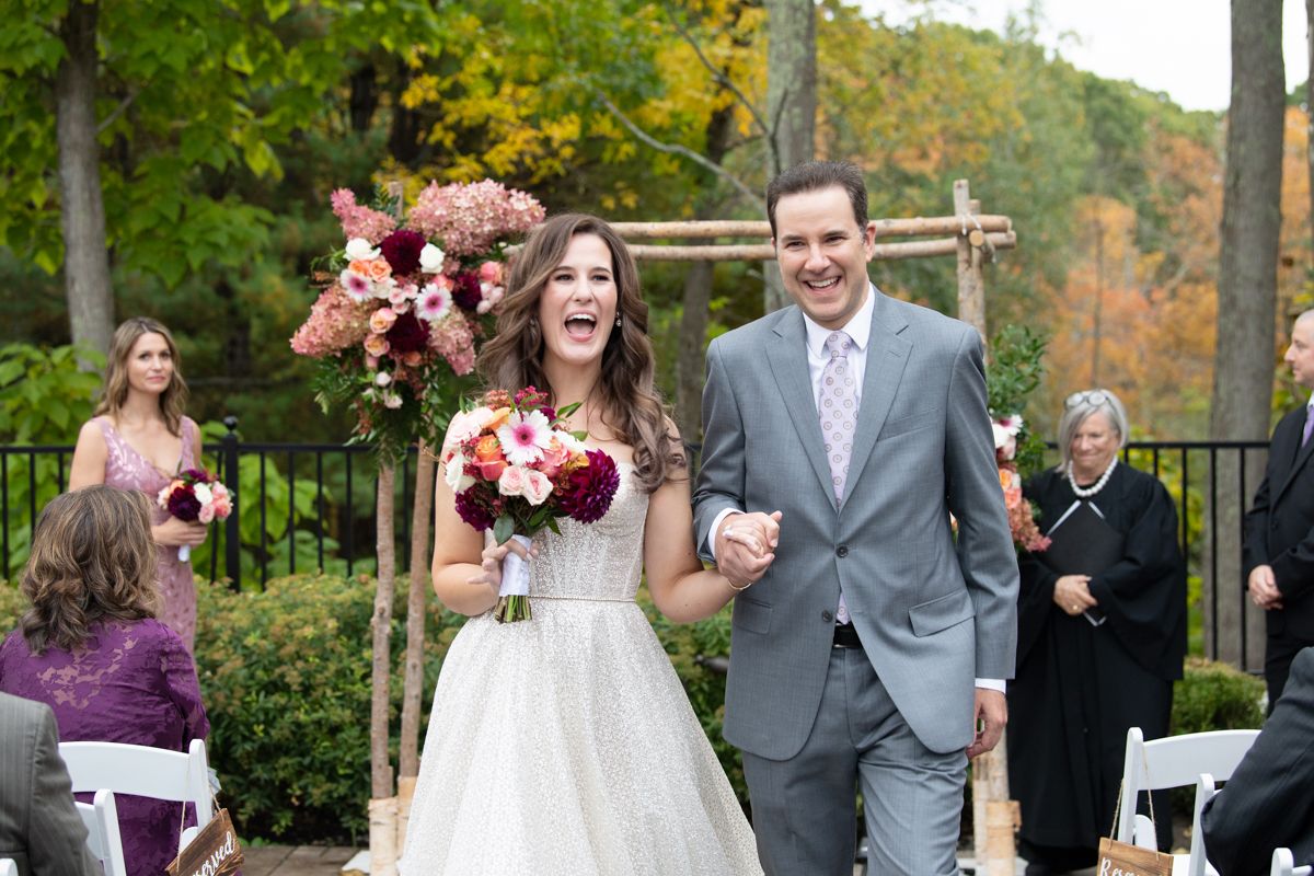 Joyful bride and groom walk back down the aisle after outdoor wedding ceremony surrounded by colorful fall foliage.