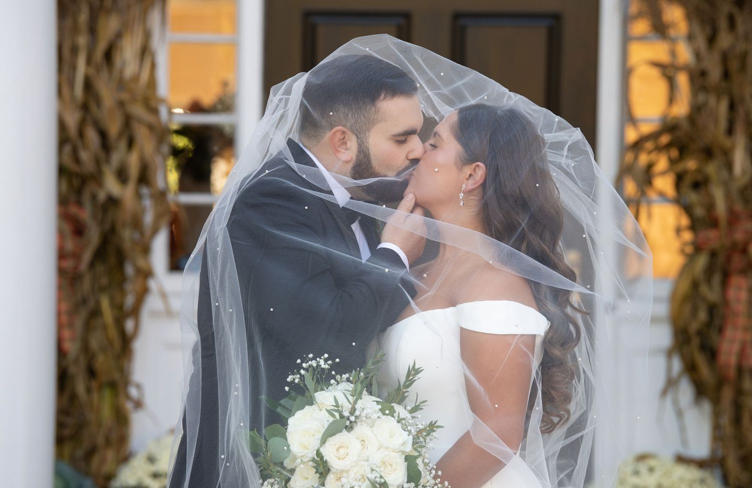 A romantic wedding couple shares a kiss under a white veil while holding a bouquet of white flowers.