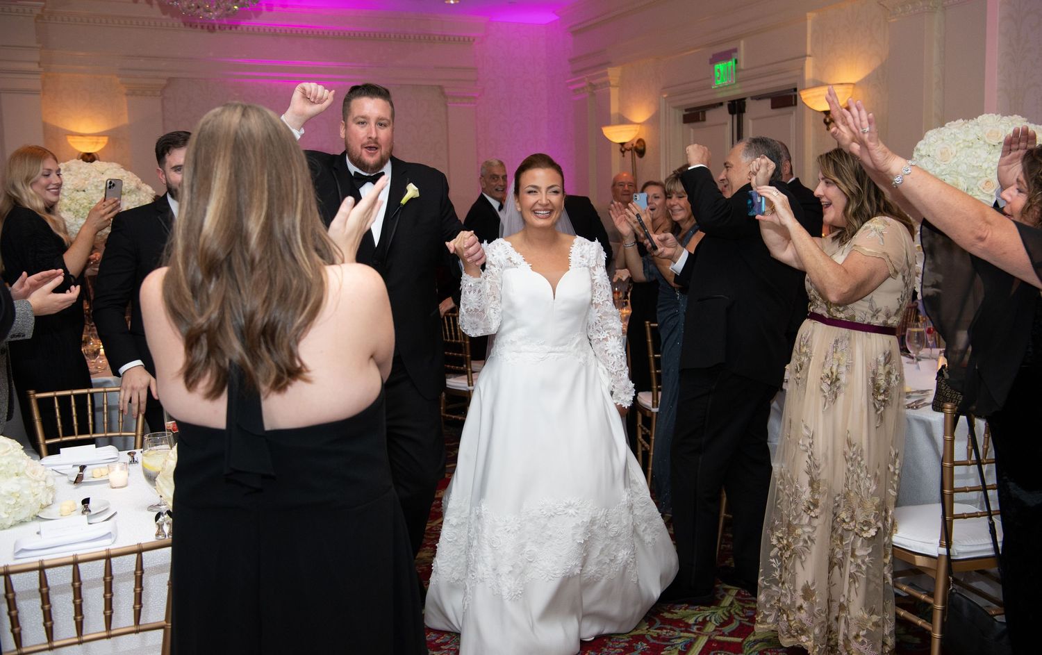 Wedding guests dance and celebrate during a reception under purple lighting in an elegant ballroom setting.