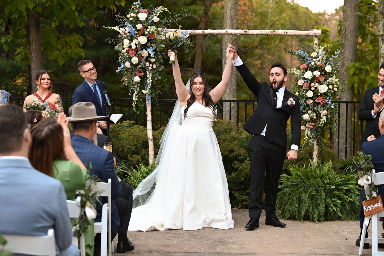 Newlyweds celebrate with raised hands under a floral arch during an outdoor garden ceremony.