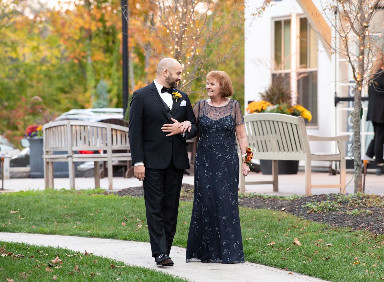 A couple in formal attire walks and chats together on a grassy lawn during autumn.