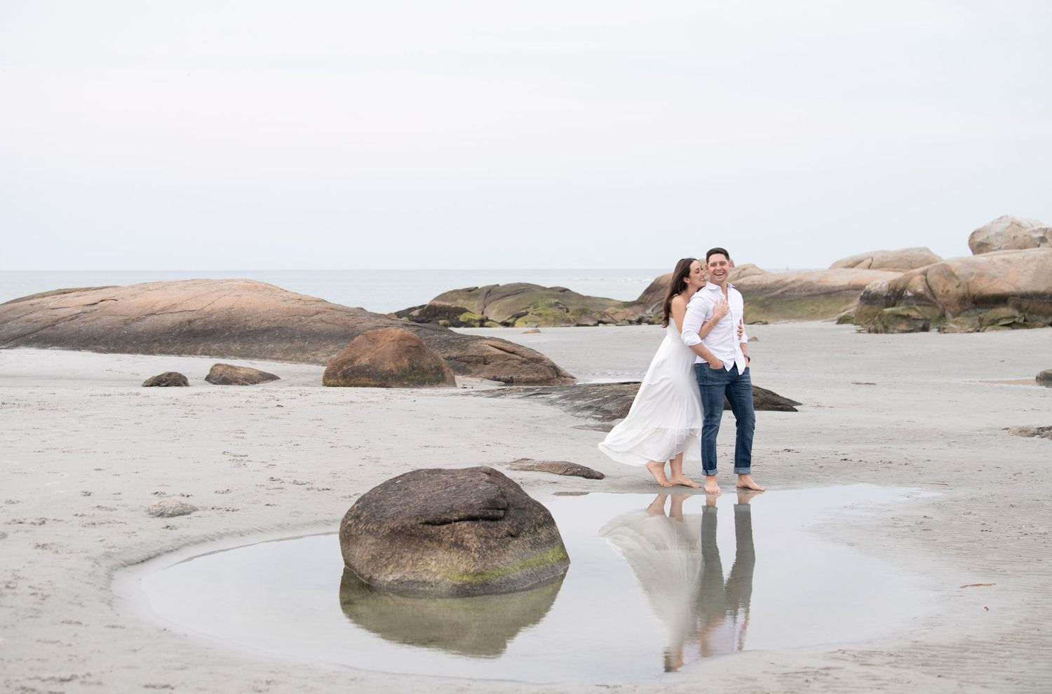 Couple embracing on rocky beach with ocean waves and sand during golden hour.