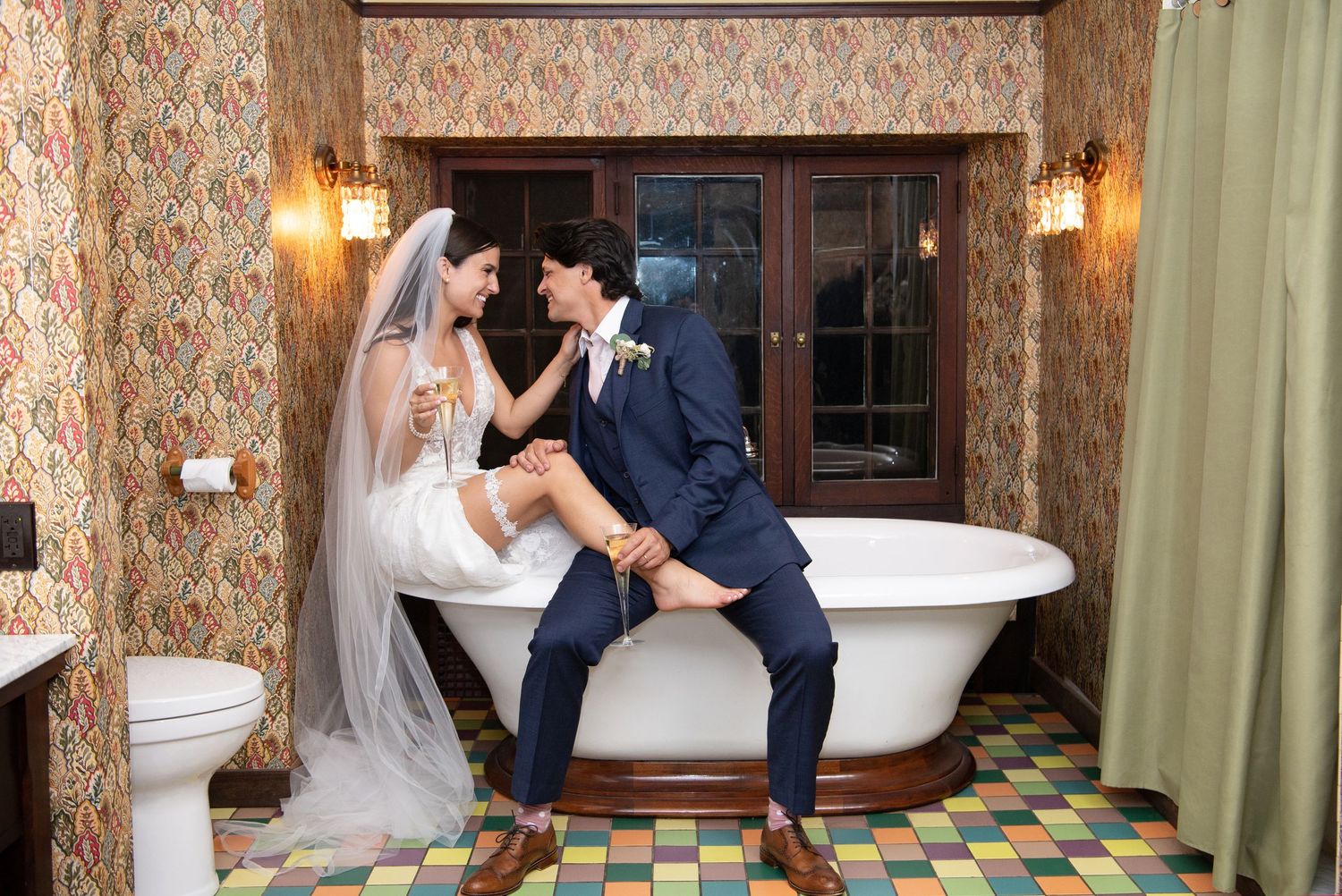 Two people share a romantic moment in a vintage bathroom with patterned floor tiles and a clawfoot tub.