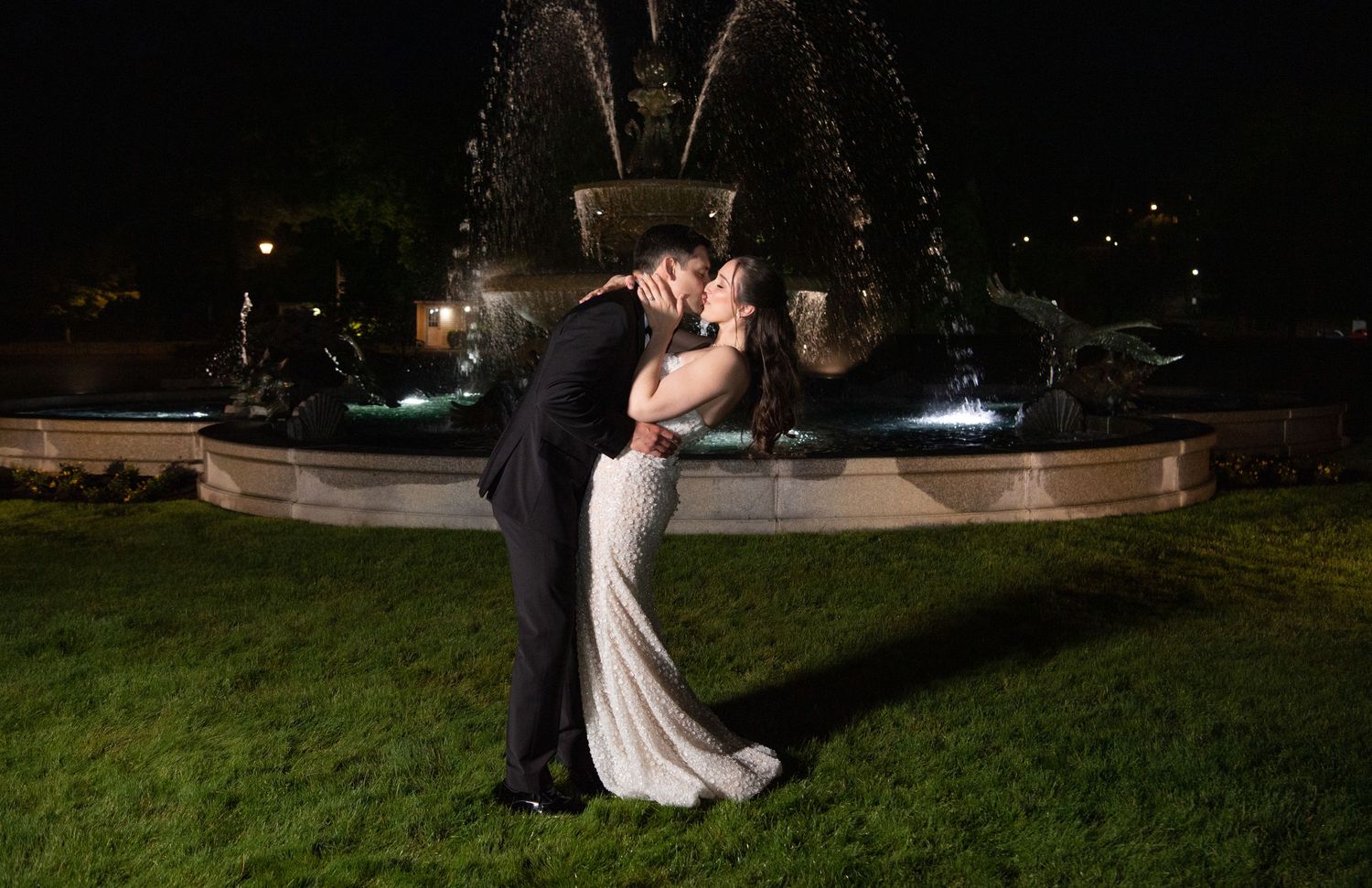 Bride and groom share a romantic kiss in front of an illuminated fountain at night during their wedding.