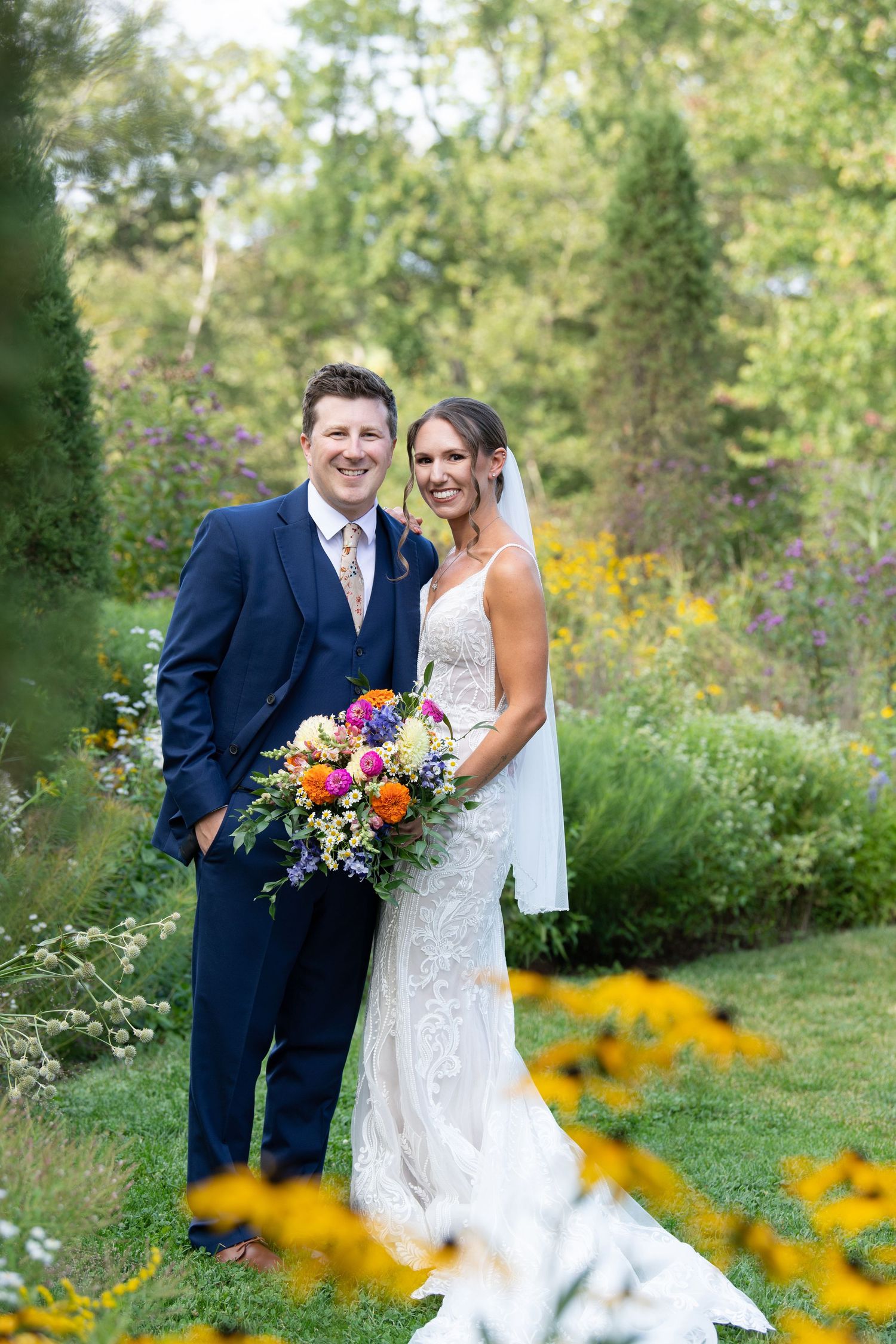 Bride and groom smiling in a lush garden, bride holding a vibrant bouquet, groom in a navy suit.