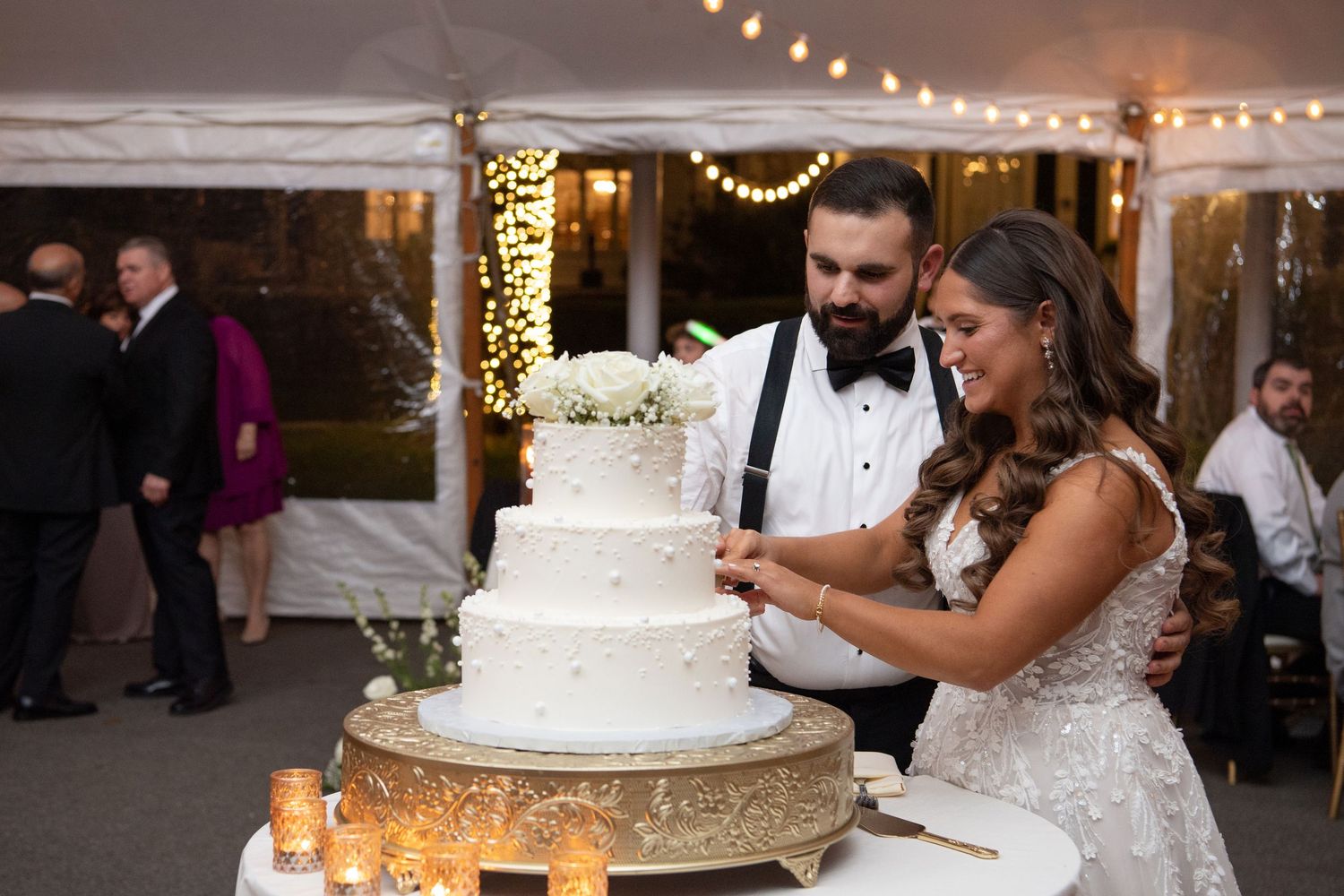Newlyweds cut their elegant white wedding cake together at a reception decorated with warm string lights.